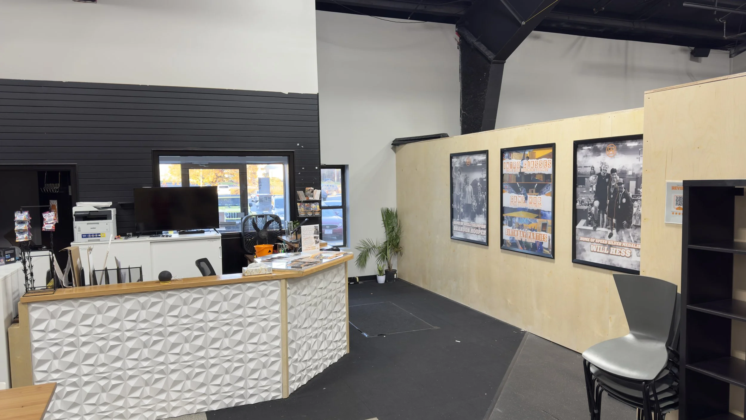 Interior of a reception or lobby area with a white textured counter, a large television, stacks of paper, and informational brochures. A black chair, black wall with horizontal paneling, a window, and a small potted plant are visible. There are three