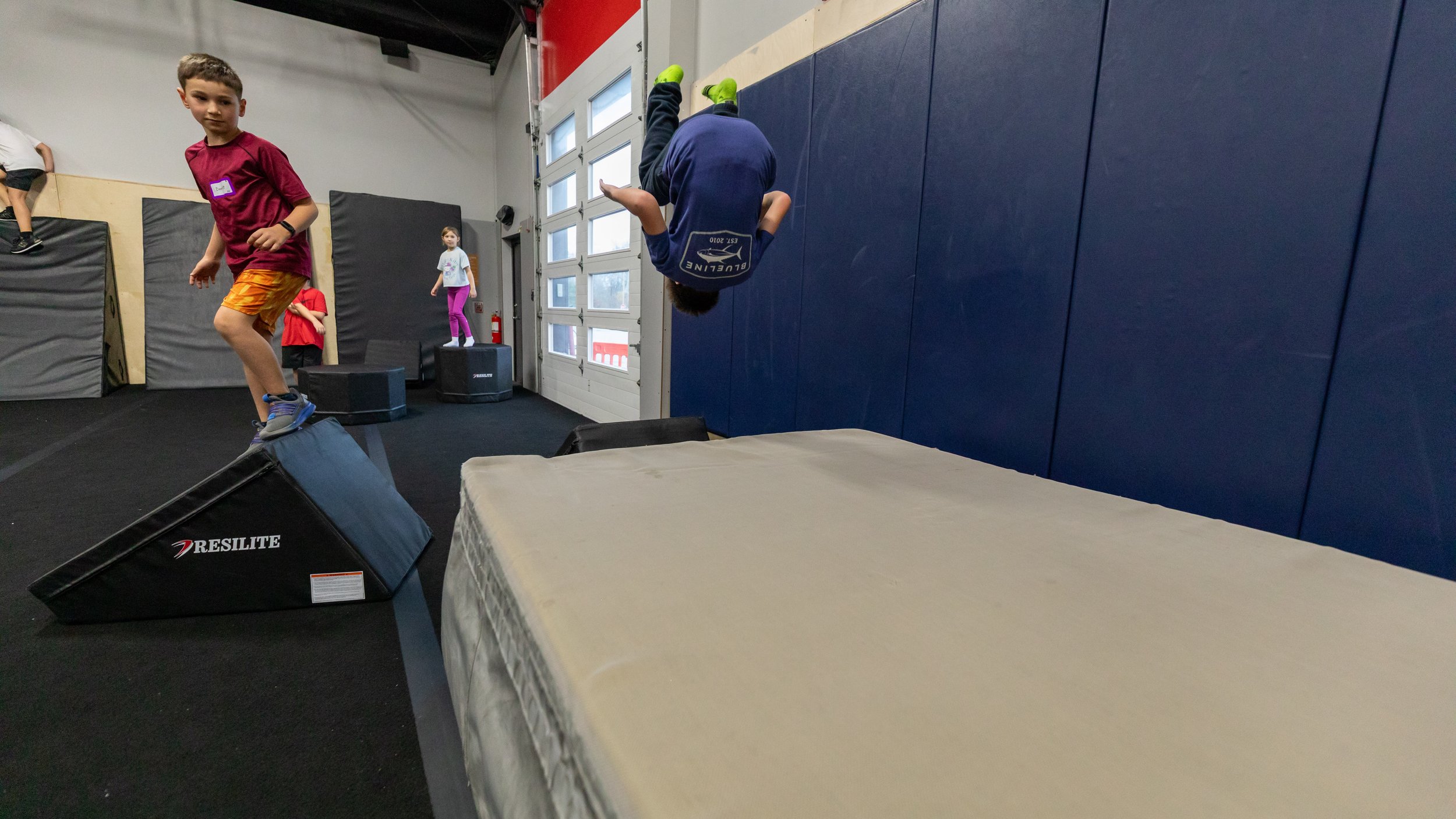 A person mid-air on a trampoline at an indoor trampoline park, with geometric mural and wooden structures in the background.