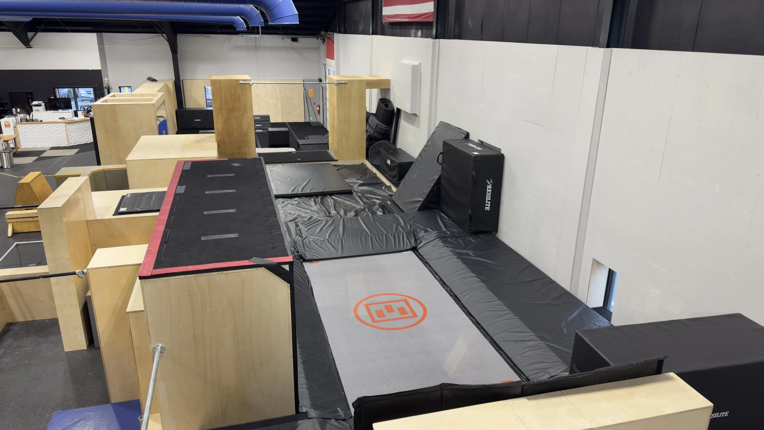 Indoor gym area with padded mats, wooden structures, and exercise equipment including a rebounder, black mats, foam rollers, and a black box, within a spacious facility with a high ceiling.
