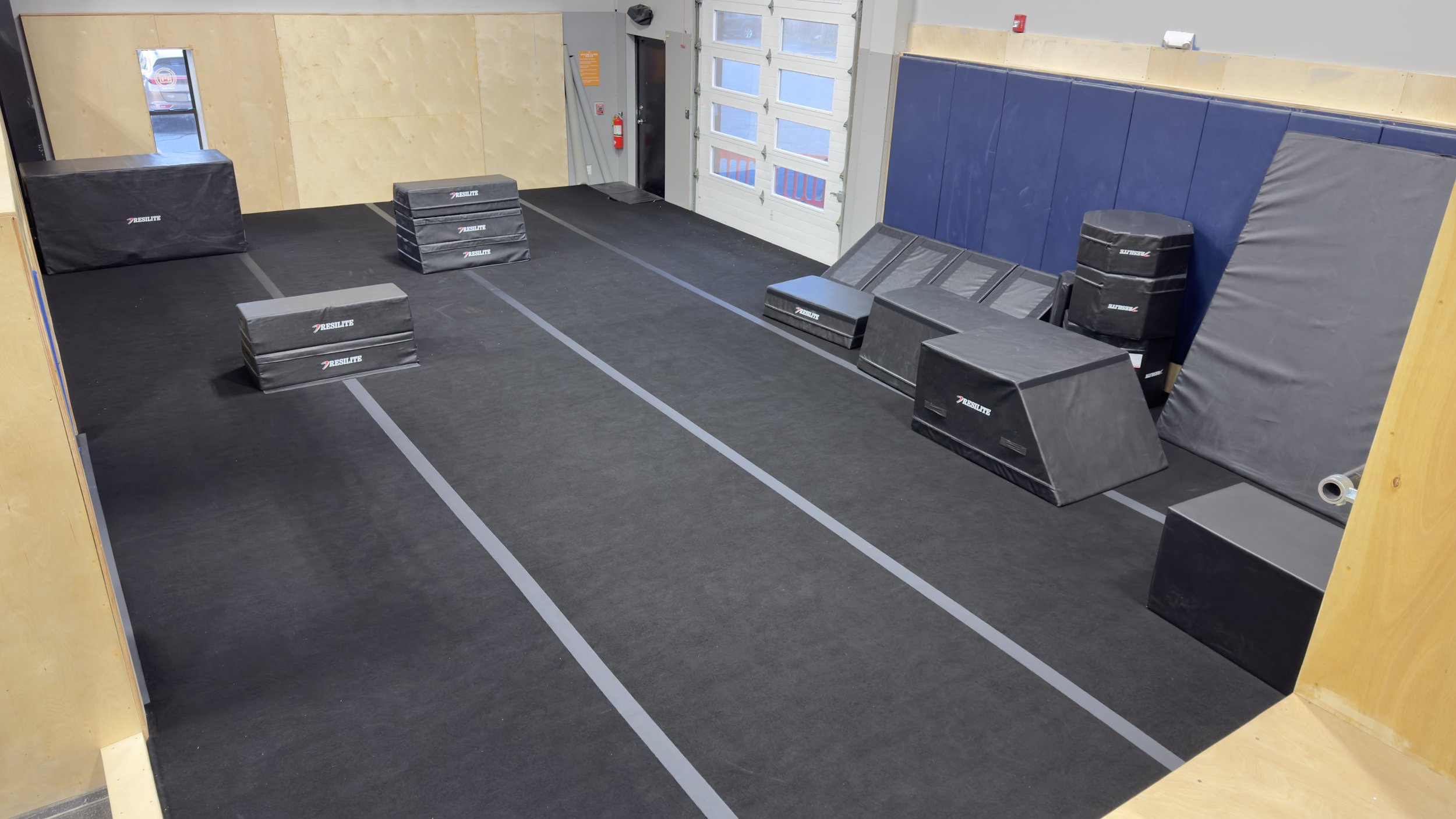 Indoor gym area with black padded workout obstacles, including boxes and ramps, arranged on a black rubber floor with marked lines, near a garage door and a blue wall.
