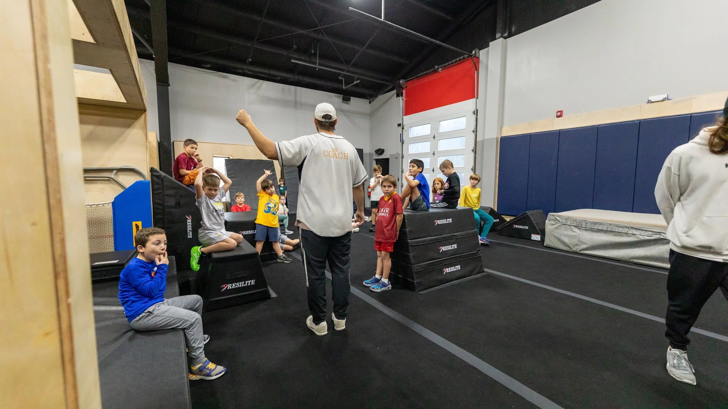 Group of children and a coach in a gymnasium, preparing for a fitness or training session, with wooden furniture and equipment in the background.