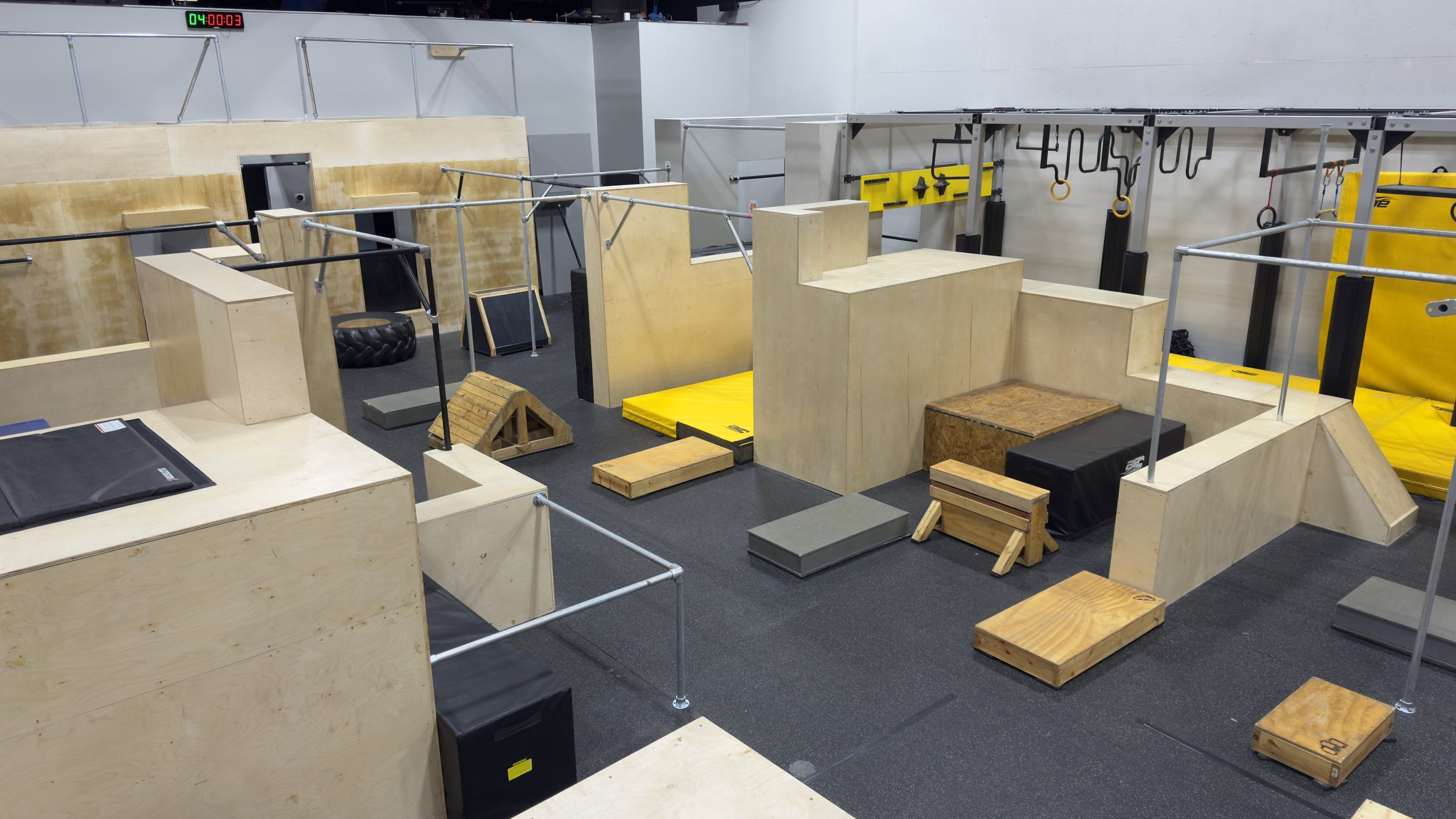 Indoor parkour training area with various wooden and padded obstacles, including walls, ramps, platforms, and climbing bars, on a rubberized floor.