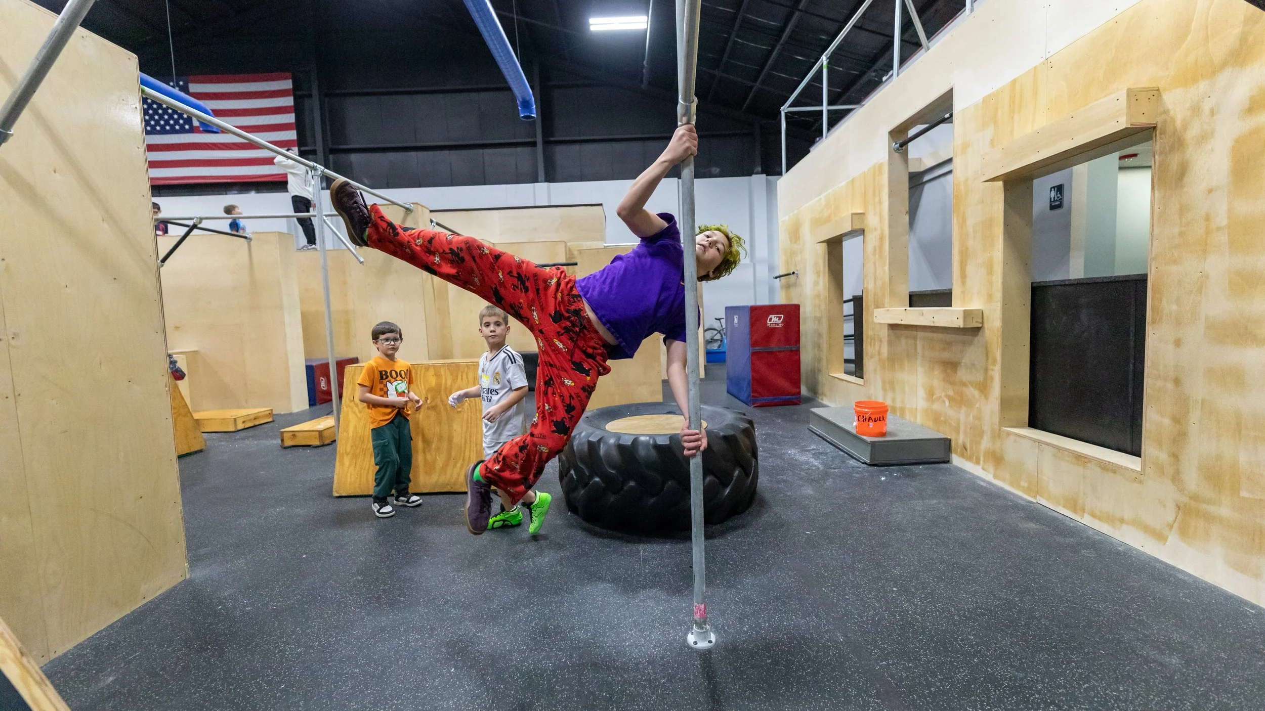 A young girl in a tie-dye shirt and purple shorts balancing on a narrow edge at an indoor trampoline park, with other kids climbing on structures in the background.