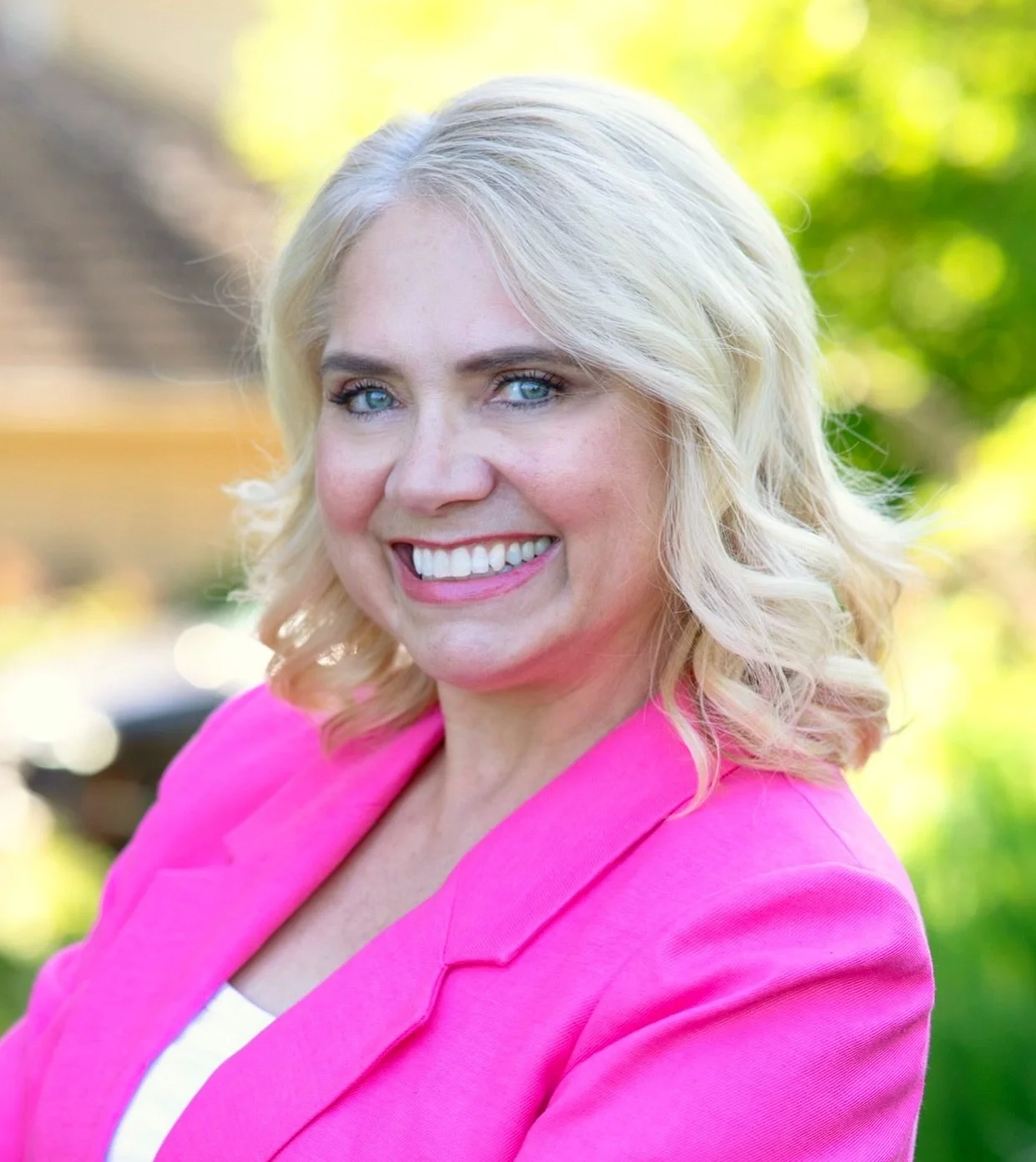 A smiling woman with blonde hair and blue eyes wearing a pink blazer outdoors with a blurred green and yellow background.