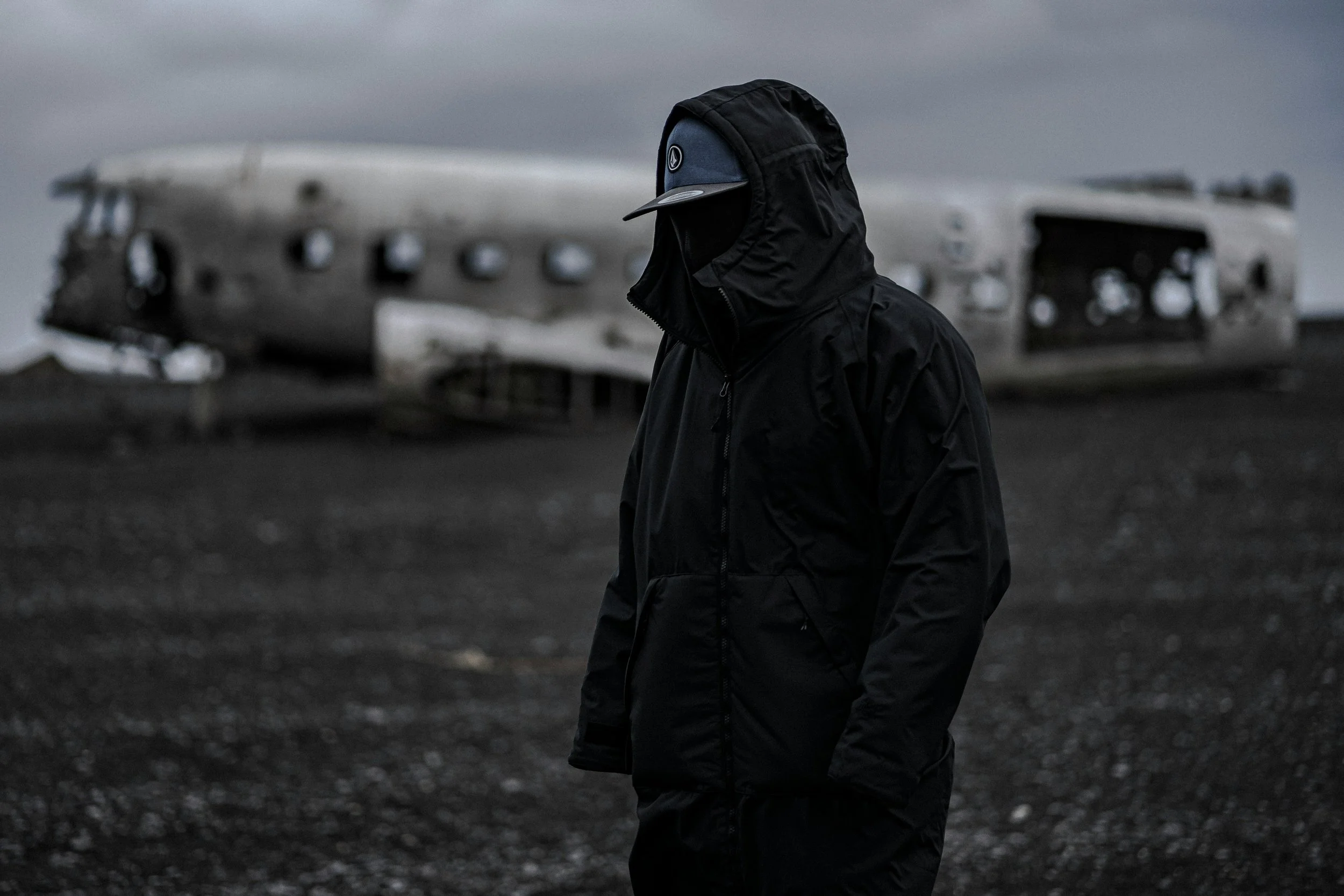a mysterious man wearing a face cover standing in front of a wrecked airplane