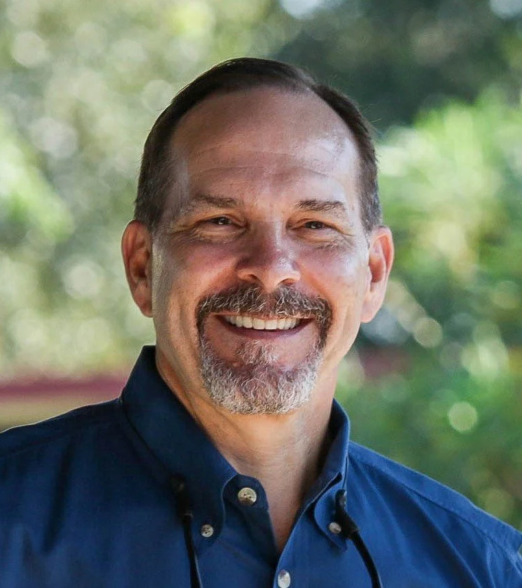 Smiling male thriller author in a blue button-down shirt, photographed outdoors with soft natural light and a blurred green background.