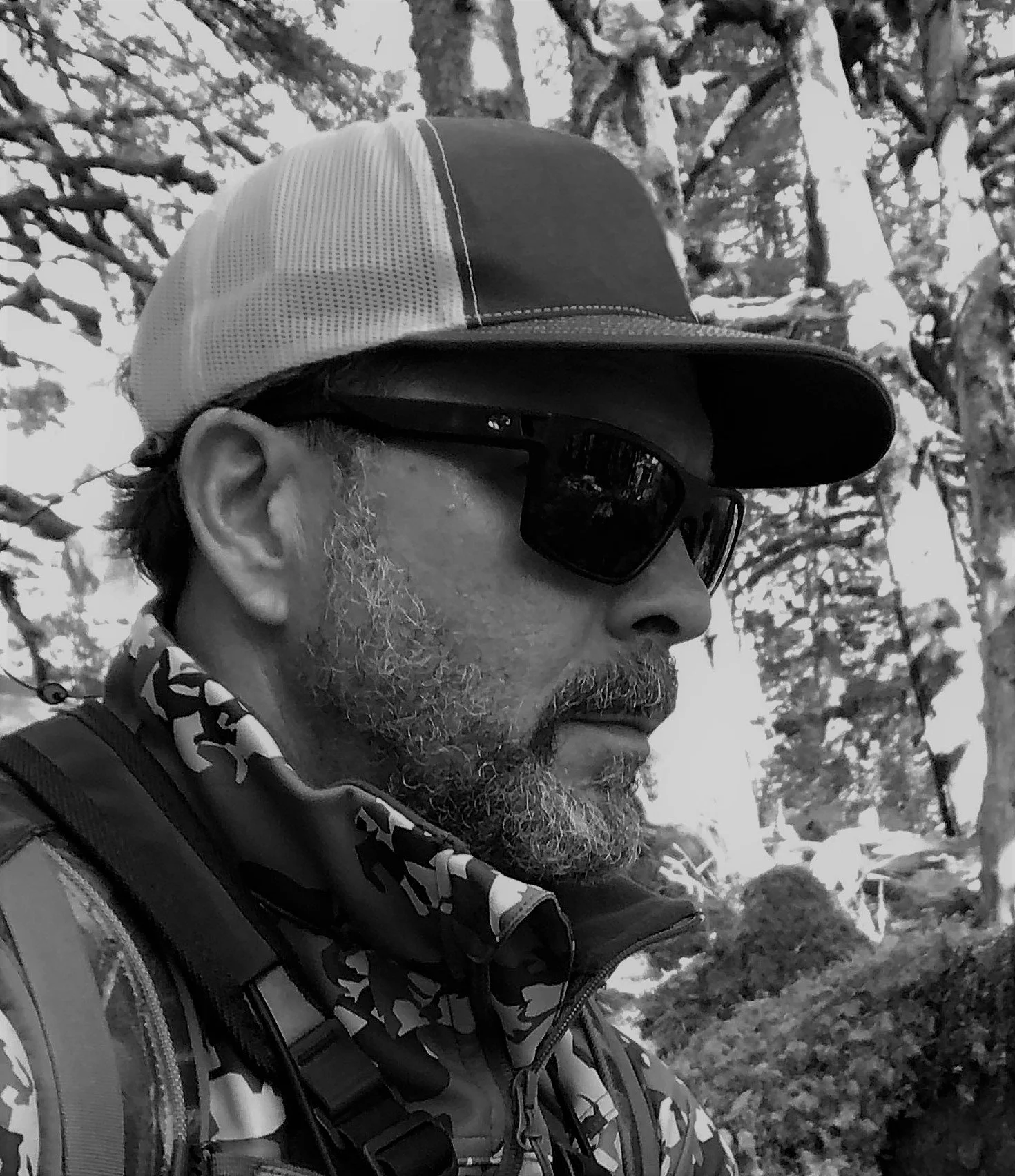 a bearded man wearing camouflage a baseball cap and sunglasses in the outdoors