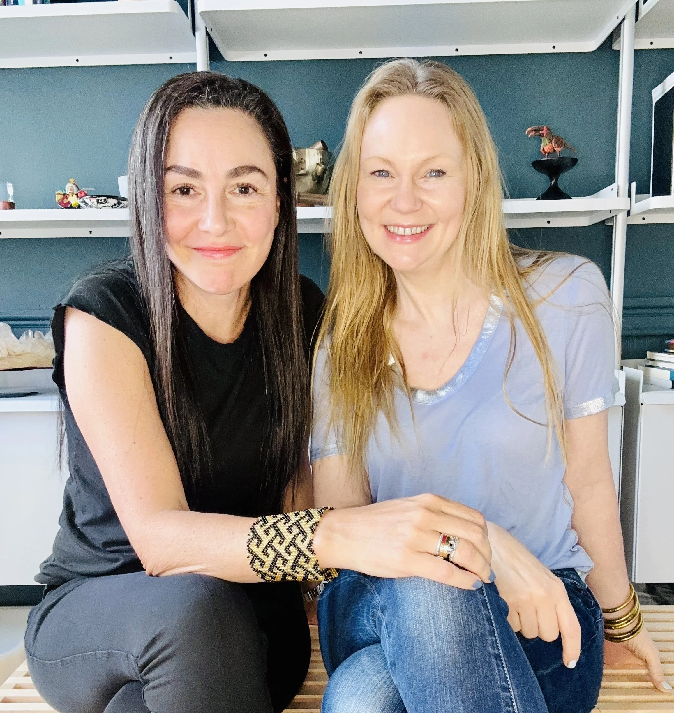 Two women sitting closely together and smiling at the camera in an indoor setting with shelves in the background.