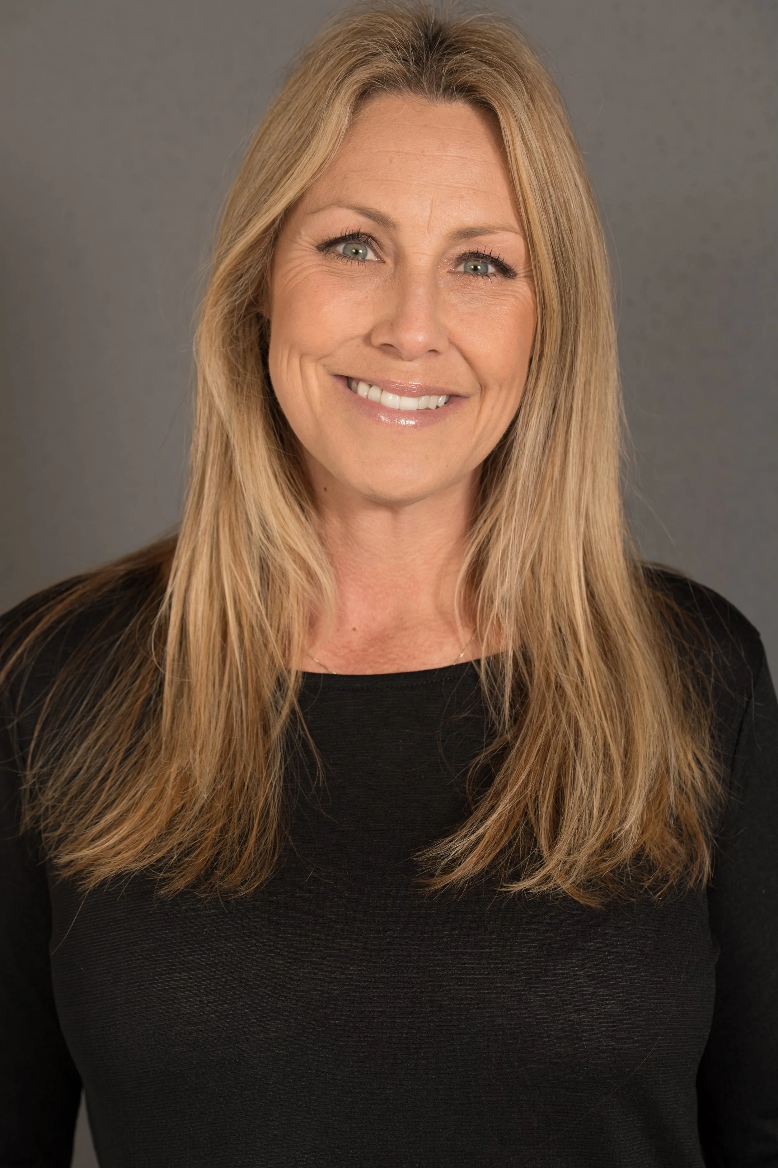 A woman with long blonde hair, wearing a black top, smiling and looking directly at the camera, against a gray background.