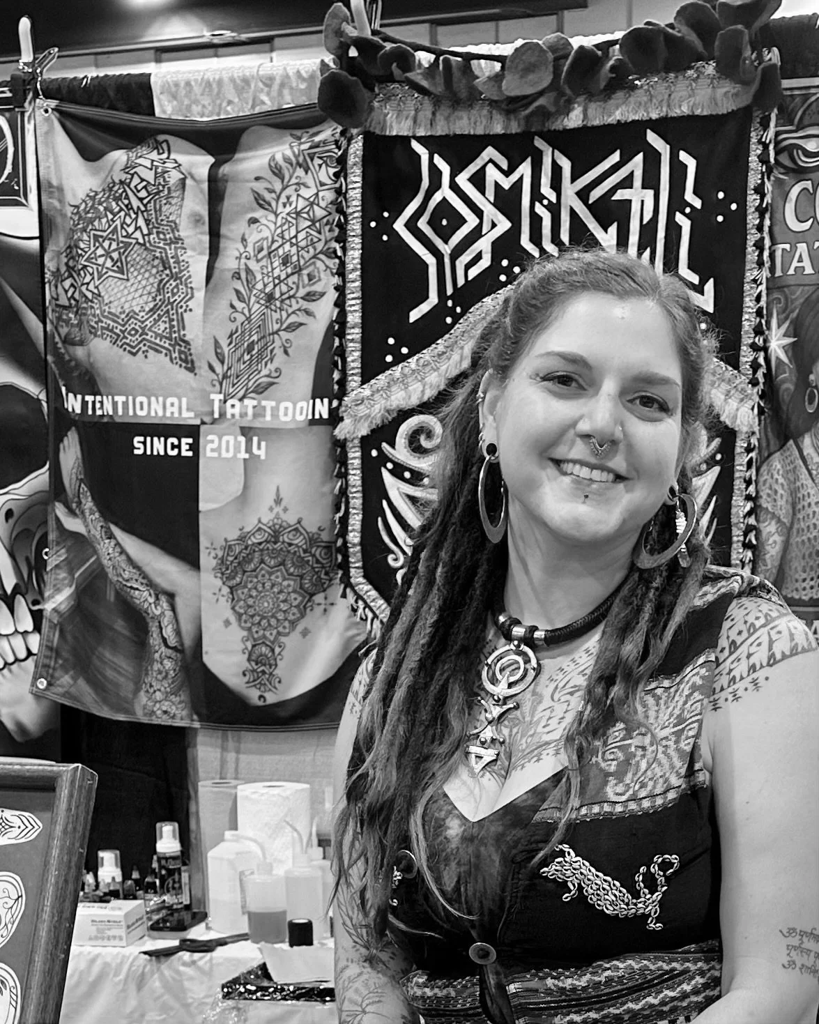 A woman with long dreadlocks, tattoos, and multiple piercings, smiling for a selfie in front of tattoo banners.