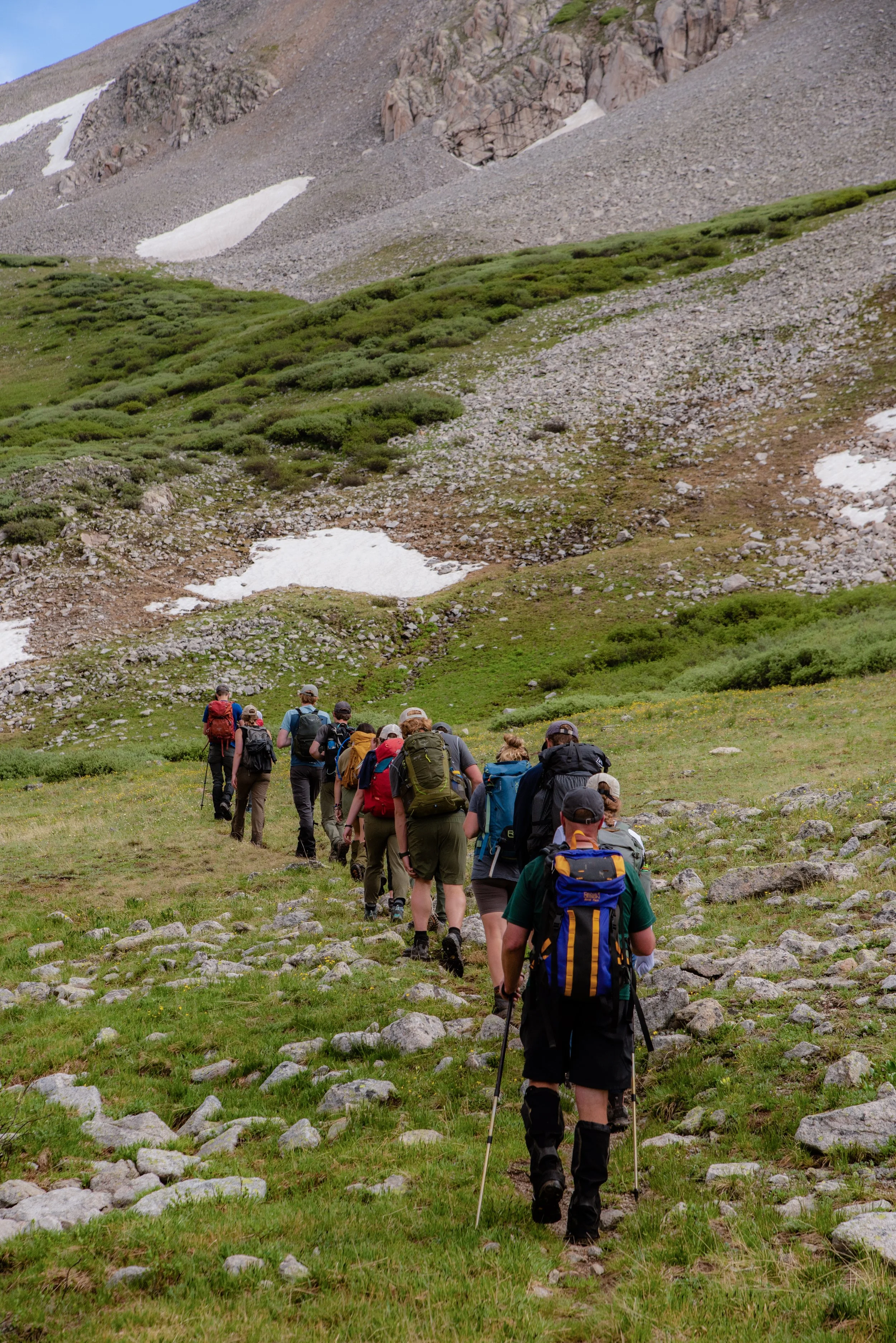 A group of hikers walking on a trail through a green mountainous landscape with patches of snow, rocky slopes, and a partly cloudy sky.