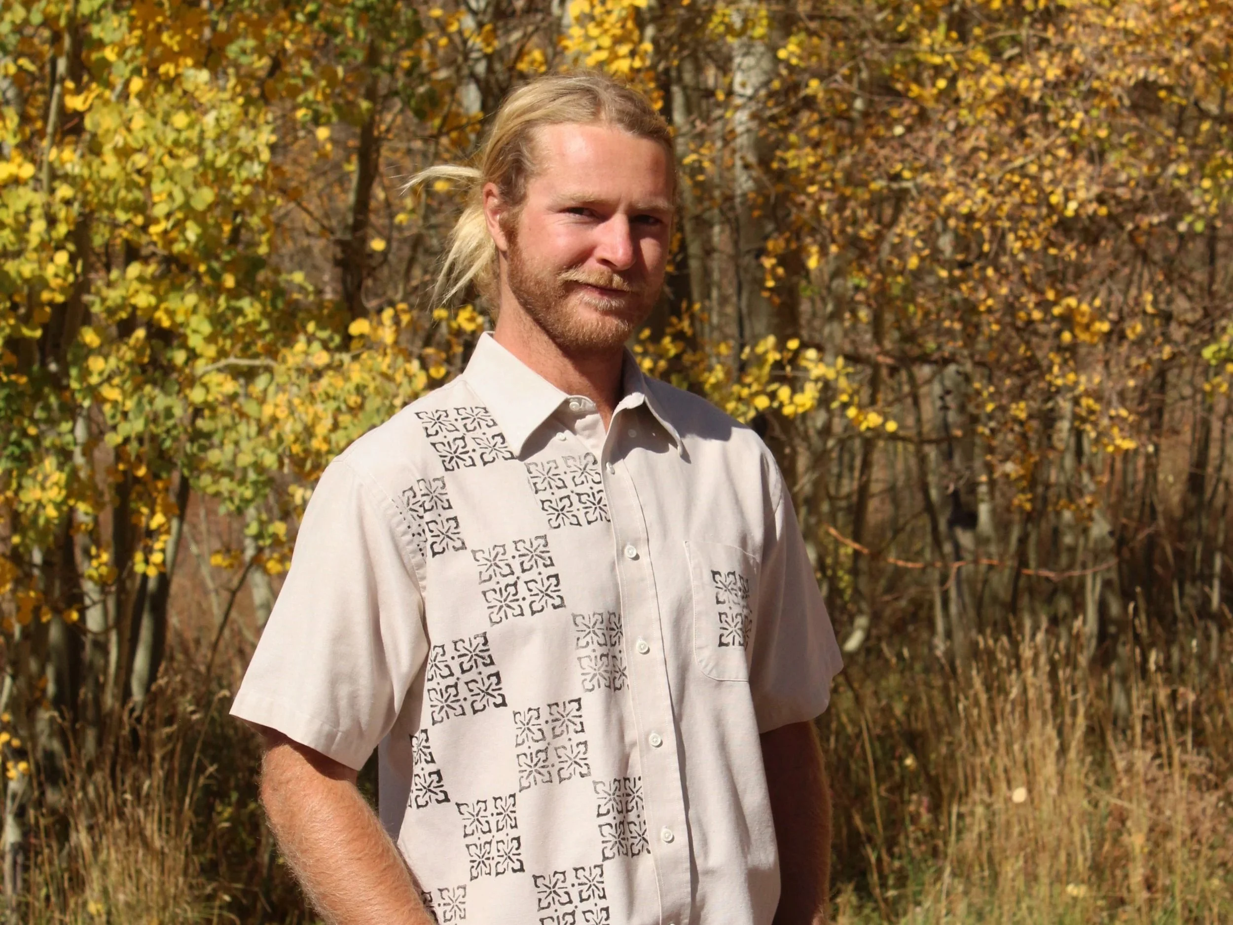 A man with long blond hair and a beard standing outdoors in autumn, wearing a light-colored shirt with decorative black patterns, in front of trees with yellow and brown leaves.