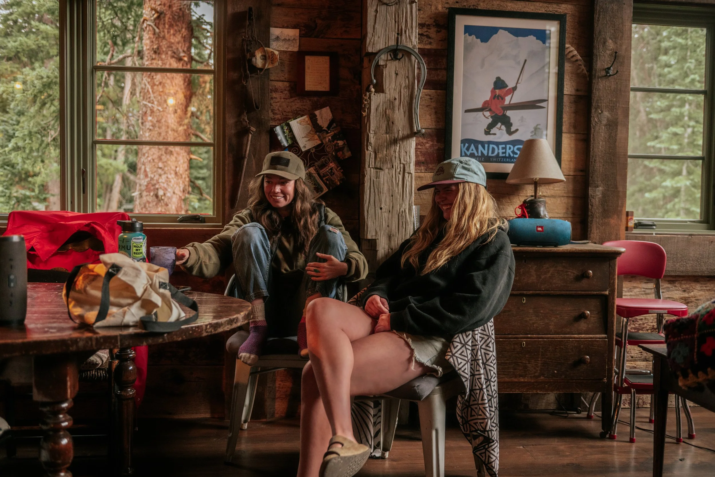 Two women sitting in a cozy rustic cabin, smiling and talking, with large windows showing tall trees outside. The woman on the left has long, wavy brown hair, is wearing a hat, a jacket, and jeans. The woman on the right has long blonde hair, is wearing a grey cap, a black jacket, and shorts. The table in front has various items, and there is a framed mountain poster on the wall.