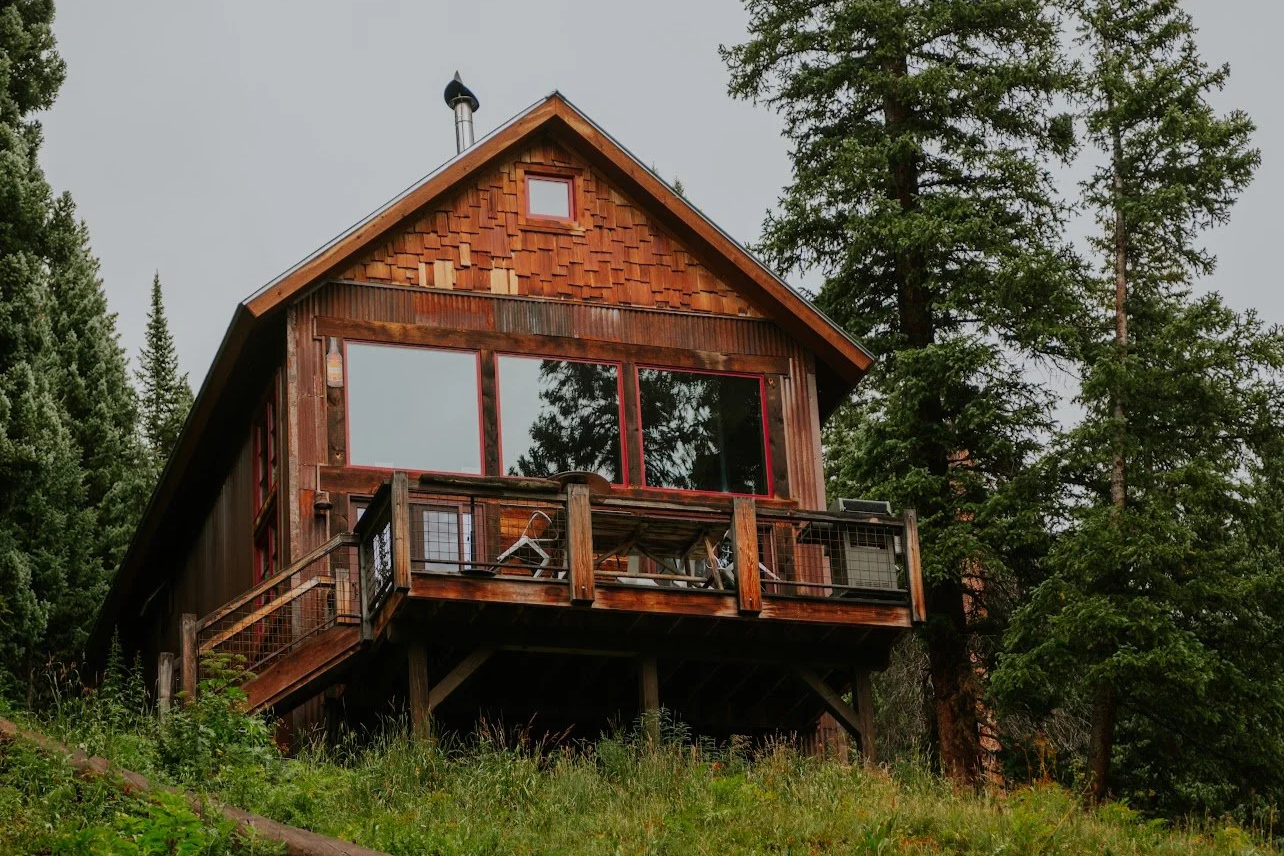 A wooden house on stilts in a forest, with a deck, railing, large windows, and a chimney on the roof.