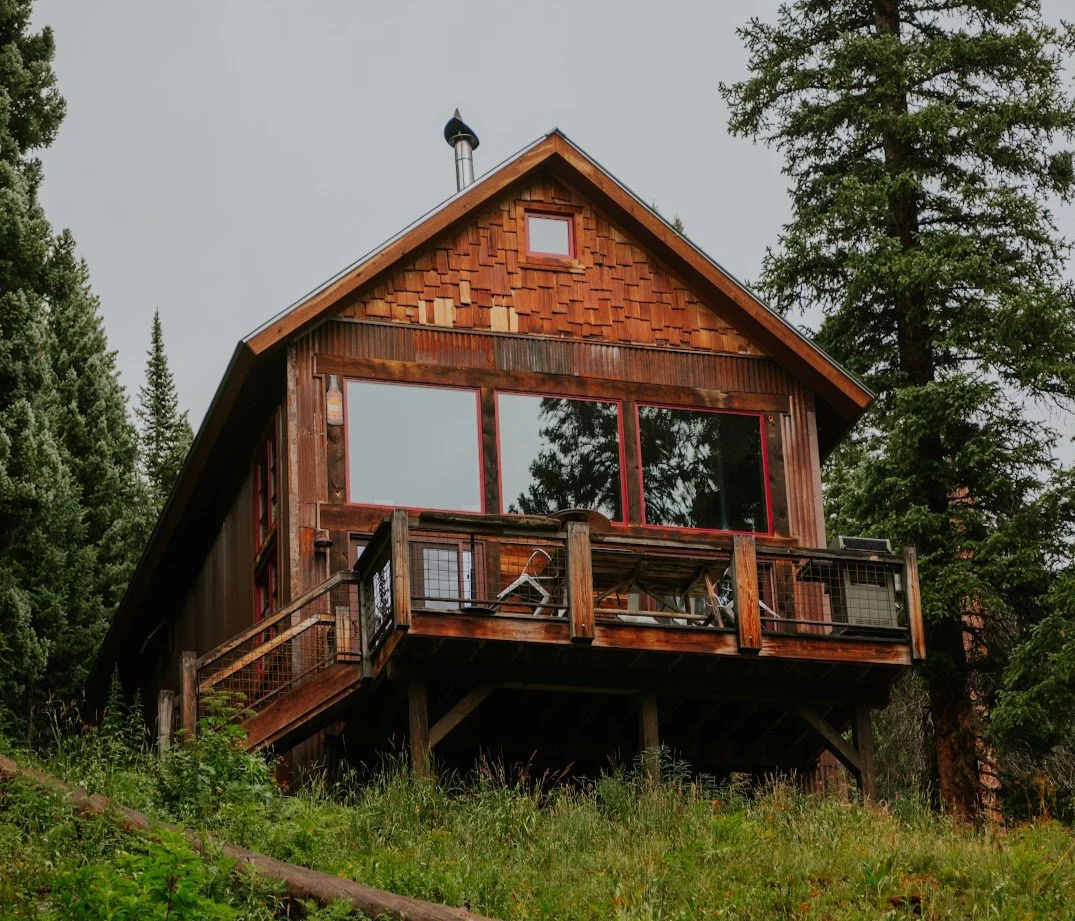 A rustic wooden house with a steep roof and large windows, situated among green trees on a hillside.