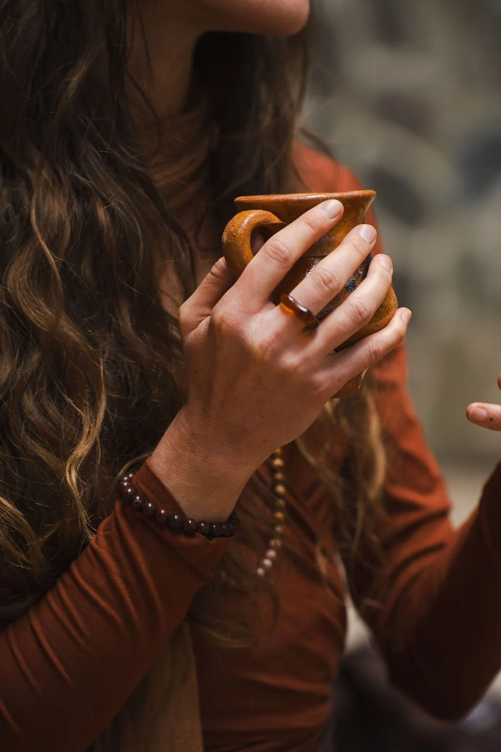 Group of women sitting cross-legged in a circle inside a cave, holding cups, participating in a ritual or ceremony, with a decorated altar in the center with flowers, candles, and offerings.