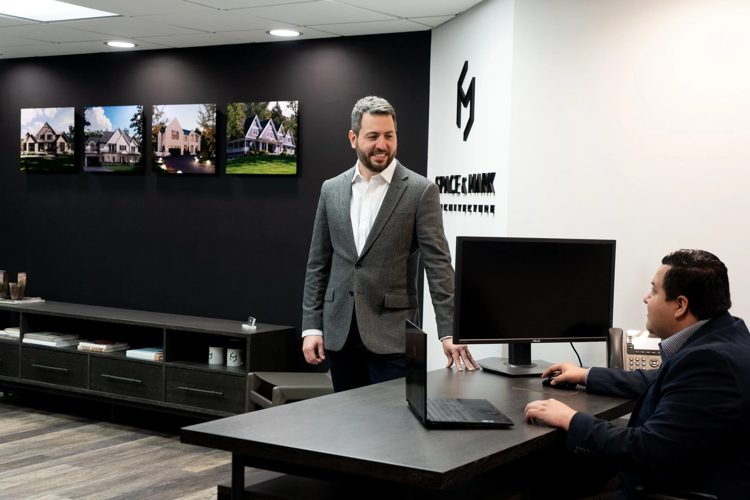 Two men in a modern office, one standing in front of a desk with a computer and the other sitting at the desk, engaging in conversation. The office features a black accent wall with framed pictures of houses, and a white wall with the company's logo.