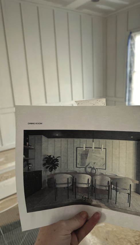 A person holding a printed interior design plan for a dining room, showing a modern space with a large artwork, a pendant light, and a row of chairs.