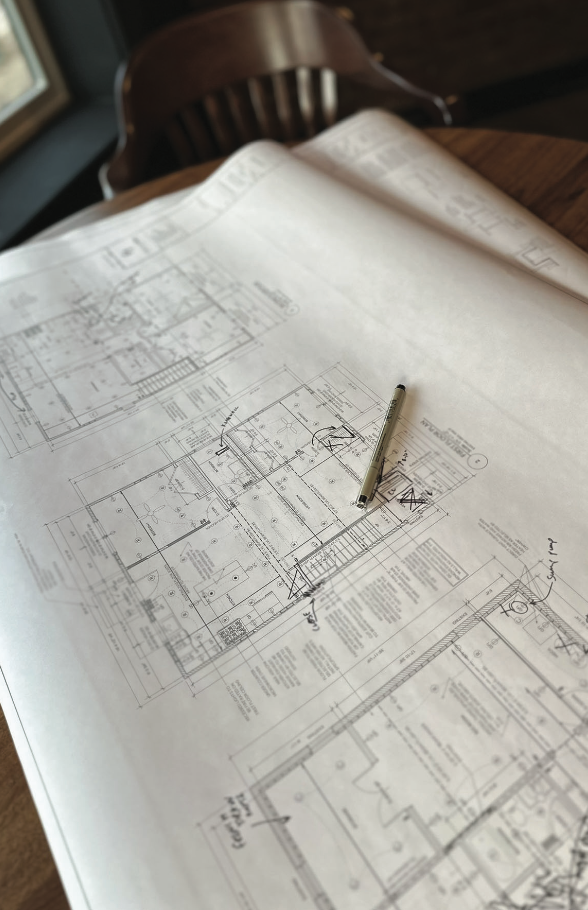 Large architectural blueprint spread out on a wooden table with a pen placed on top, near a wooden chair in a room with natural light coming through a window.