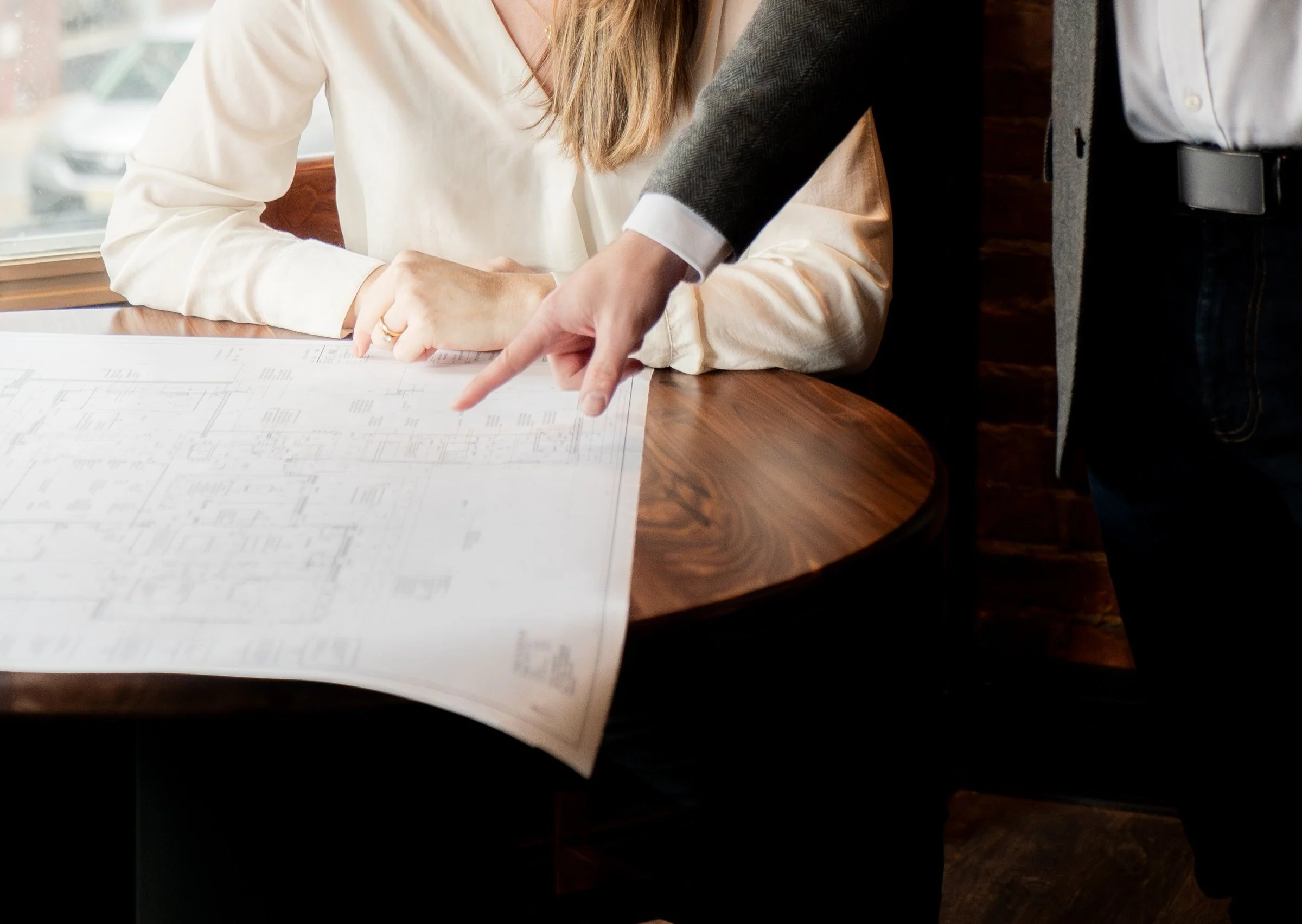 A person in a suit points at a blueprint on a wooden table while a woman in a white shirt looks on in a room near a window.