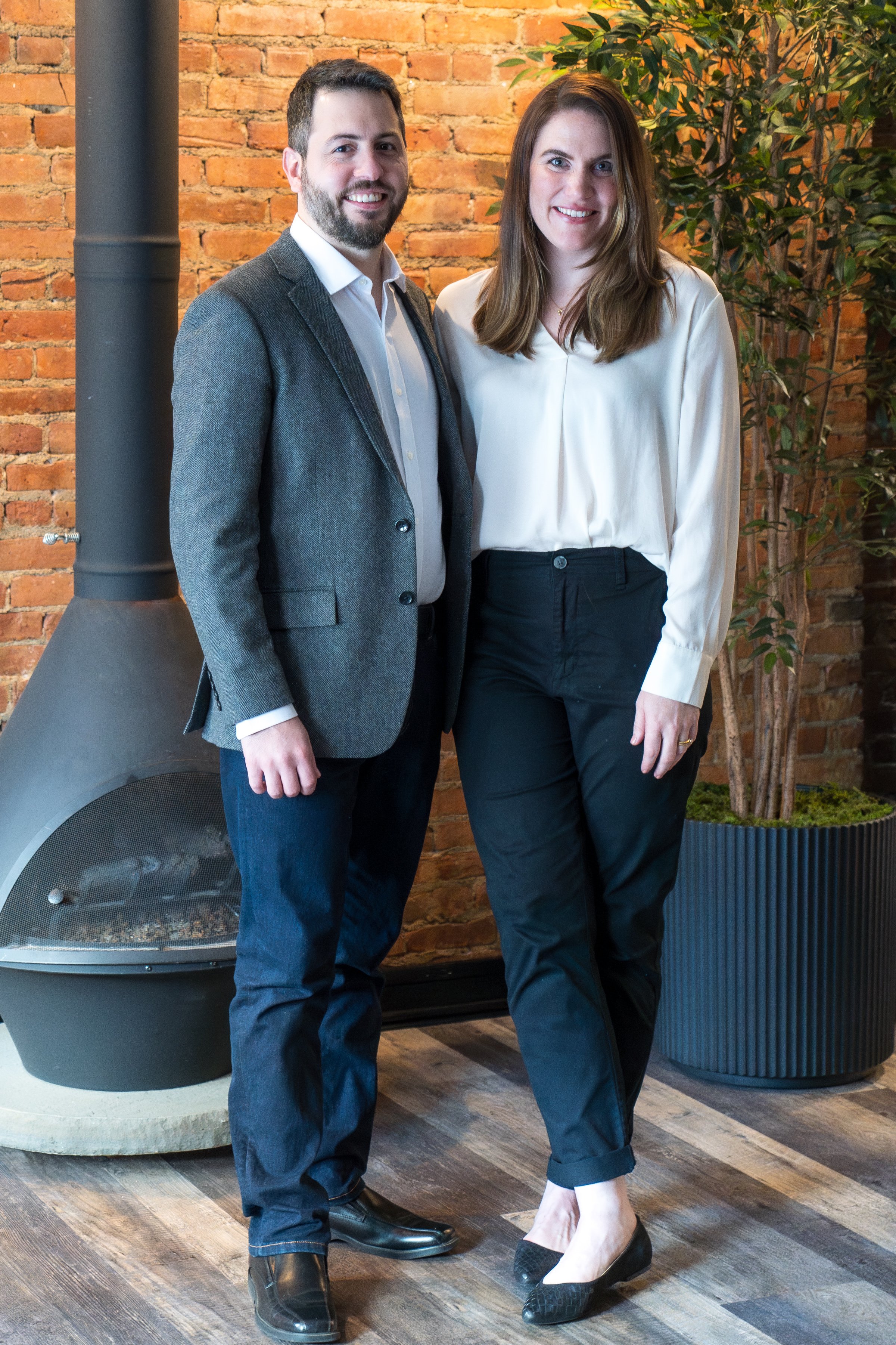A man and woman standing together indoors, smiling, in front of a brick wall and a large potted plant.