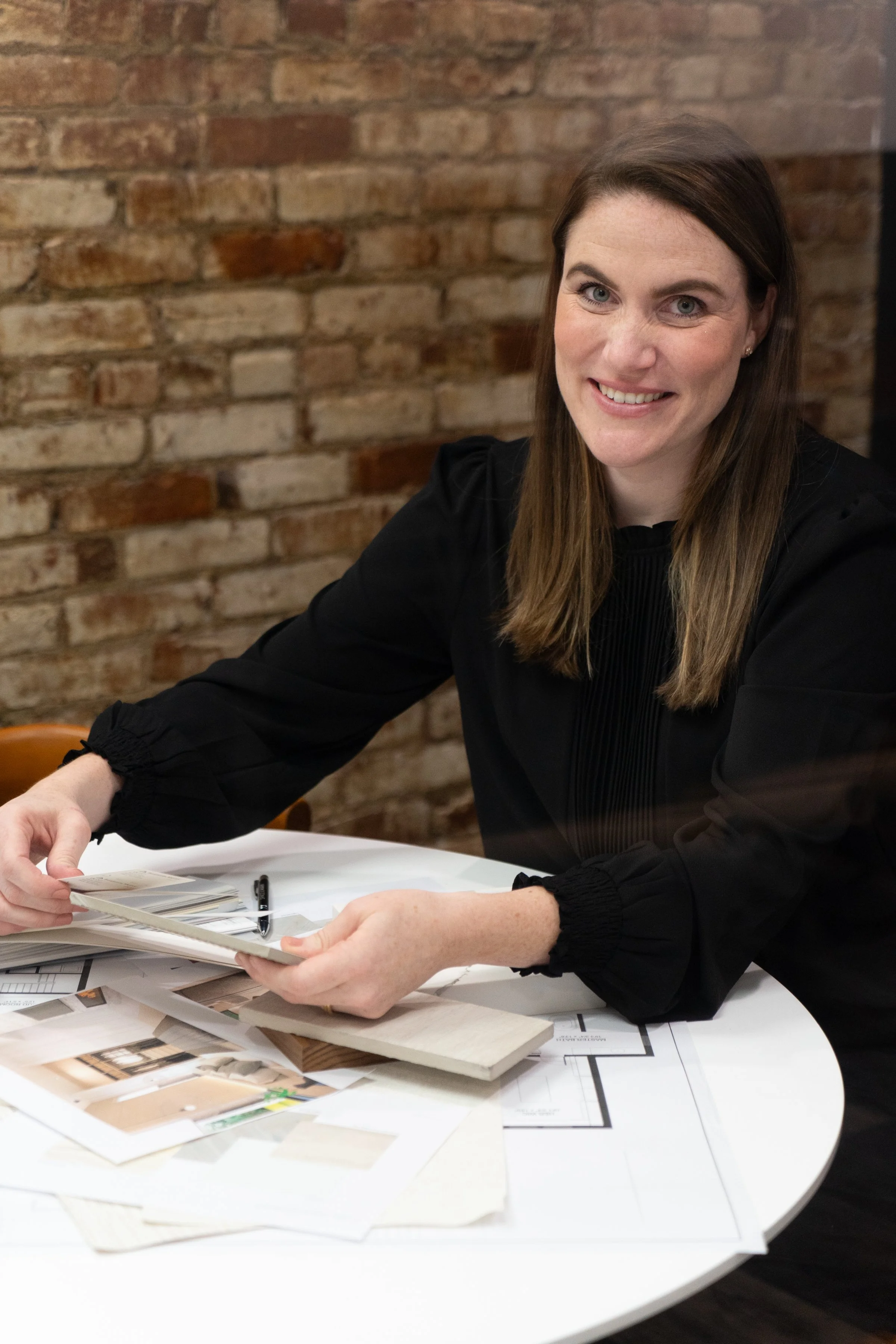A woman sitting at a table, looking at the camera, with materials and photos spread out in front of her, and a brick wall in the background.