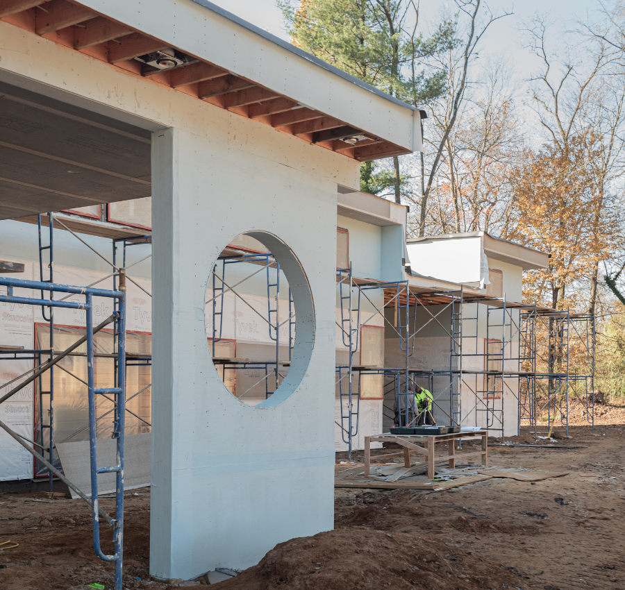 Construction site with scaffolding around a building under construction, trees in the background, and a worker in a yellow vest working on the structure.