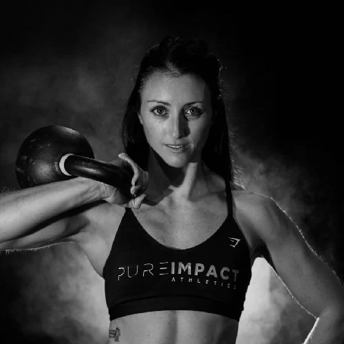 Amber Tait fit woman with long dark hair in athletic wear holding a kettlebell on her shoulder in a gym. Black and white photograph with smoke in the background.