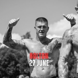 A shirtless man with tattoos flexing his arms outdoors, with the words 'Bolton 27 June' in red and white text overlay.