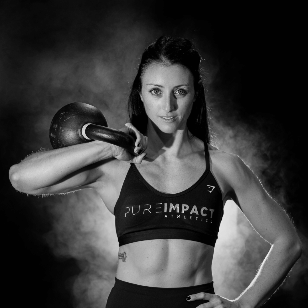 A fit woman with long dark hair in athletic wear holding a kettlebell on her shoulder in a gym. Black and white photograph with smoke in the background.