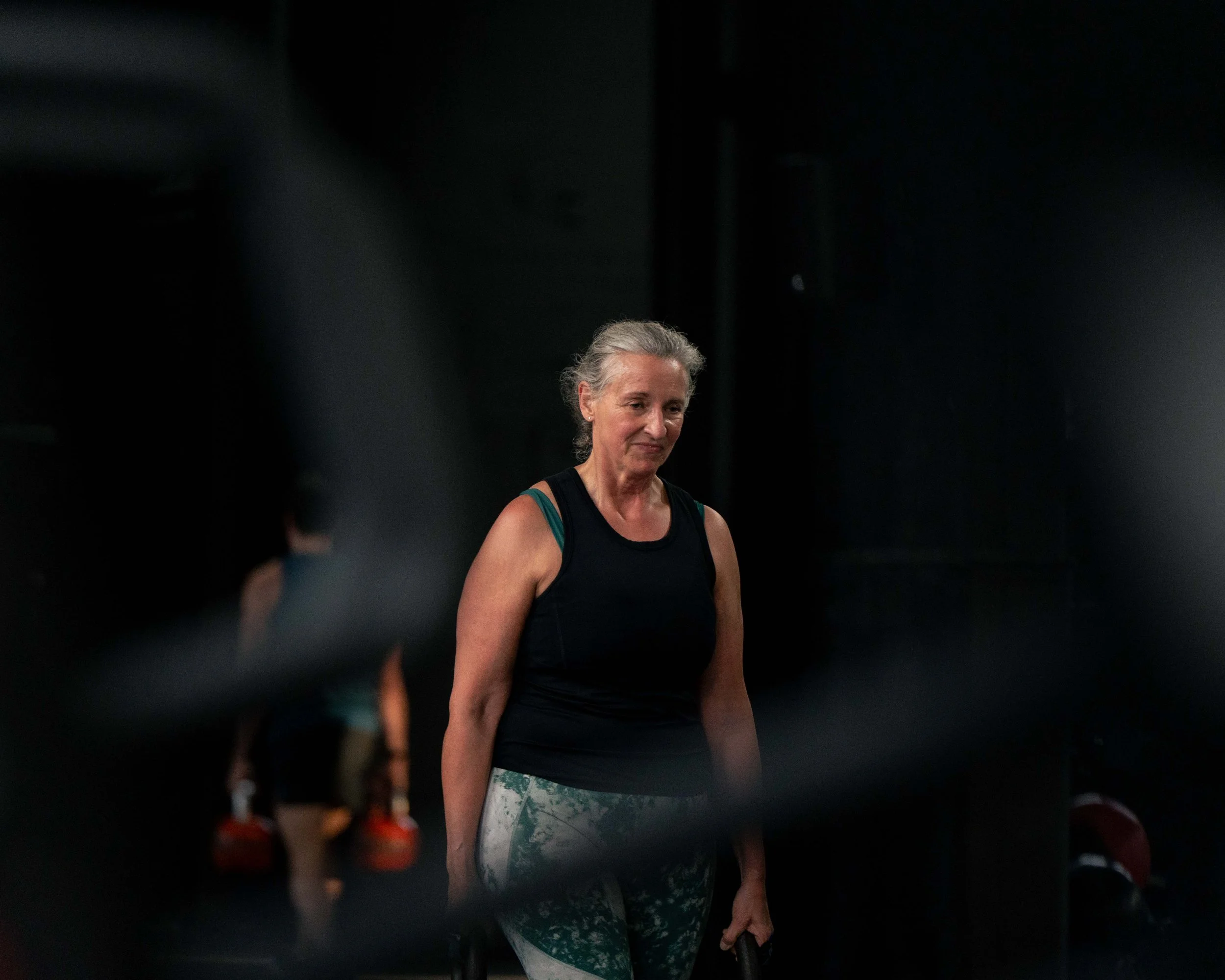 An older woman with gray hair in a ponytail, wearing a black tank top and patterned leggings, standing in a gym.