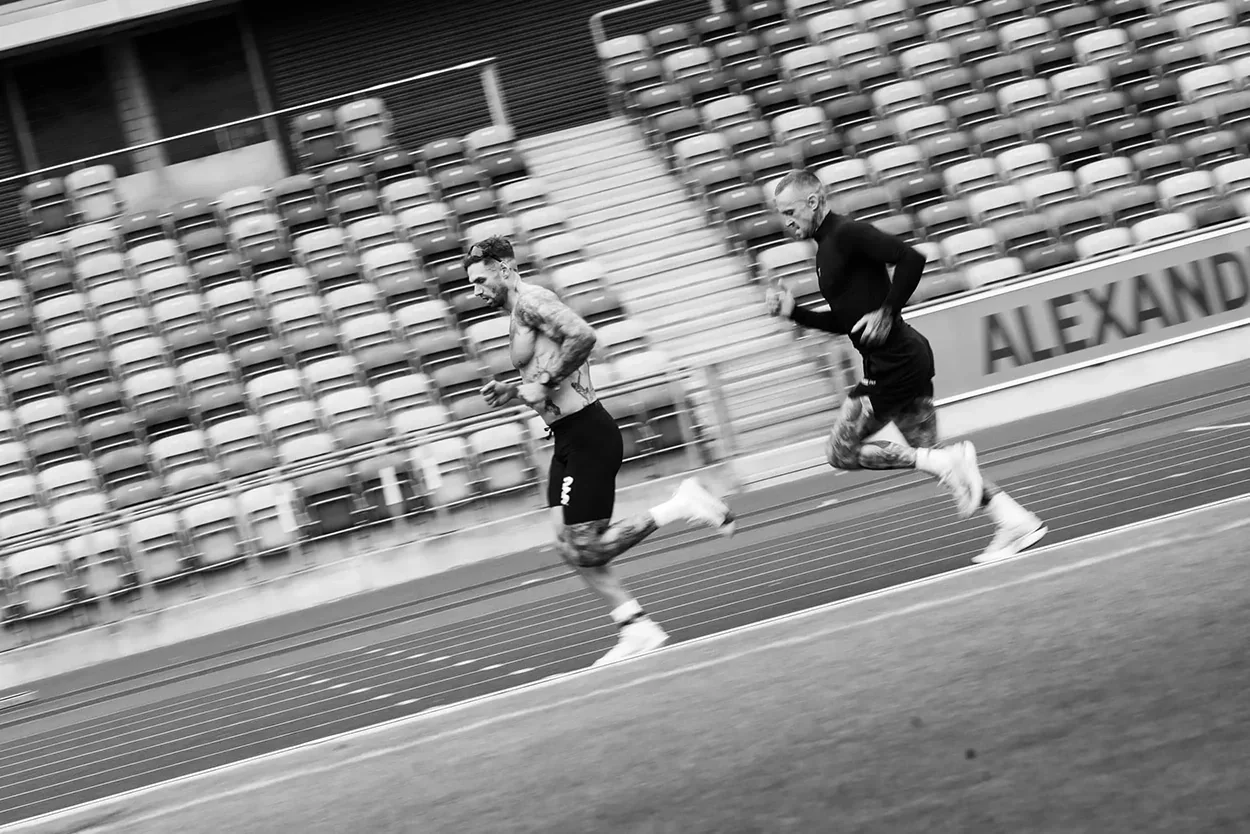 Two tattooed men running on a track inside a stadium with empty seats in the background.