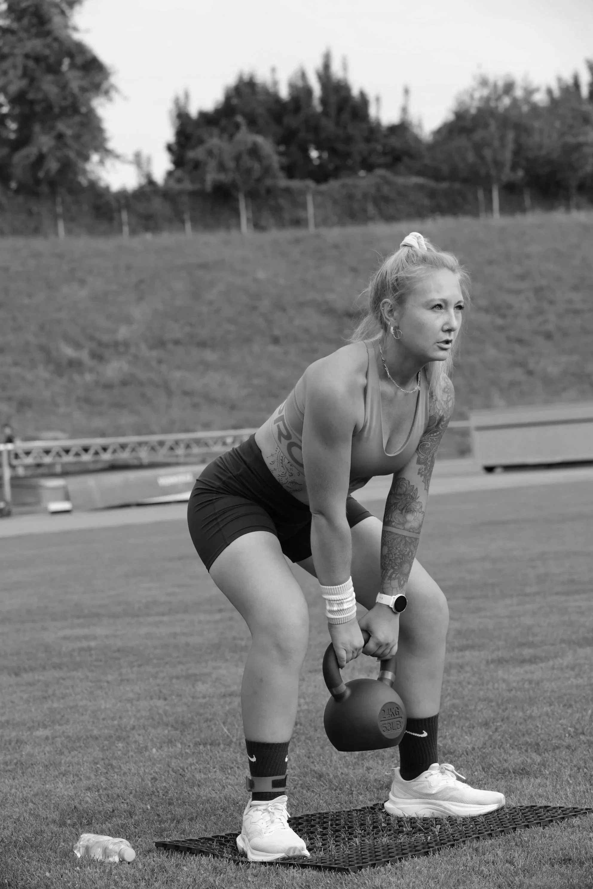 A woman with tattoos, wearing athletic gear and a watch, is bent over, gripping a kettlebell with both hands outdoors on a grassy field, preparing for exercise. There's a water bottle on the ground nearby.