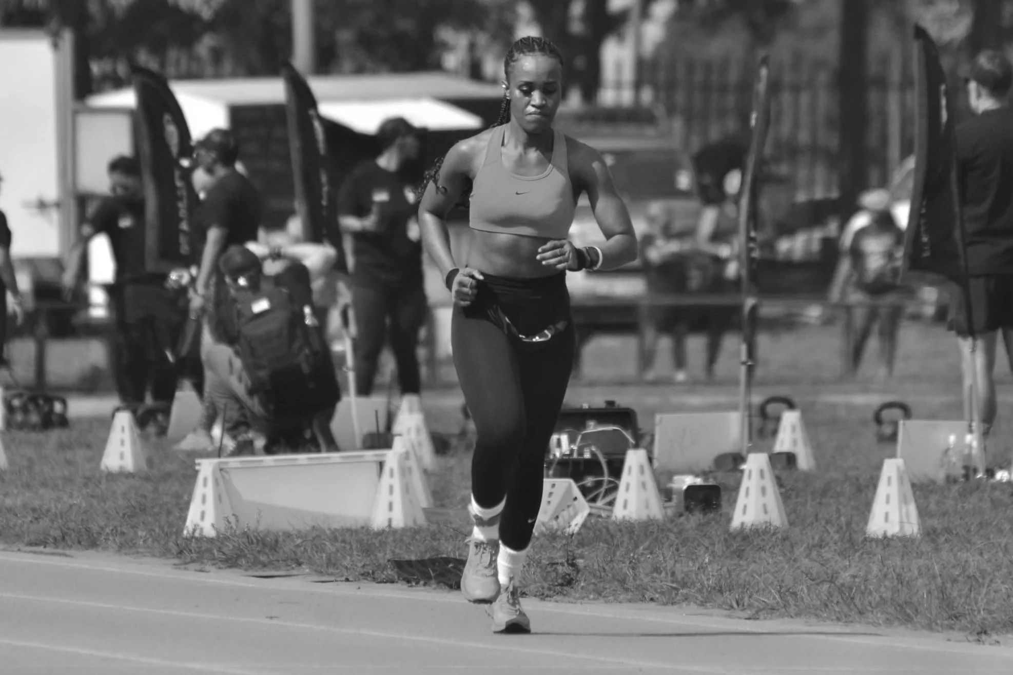 A woman running outdoors during a track event, wearing athletic gear, with other people and equipment in the background.