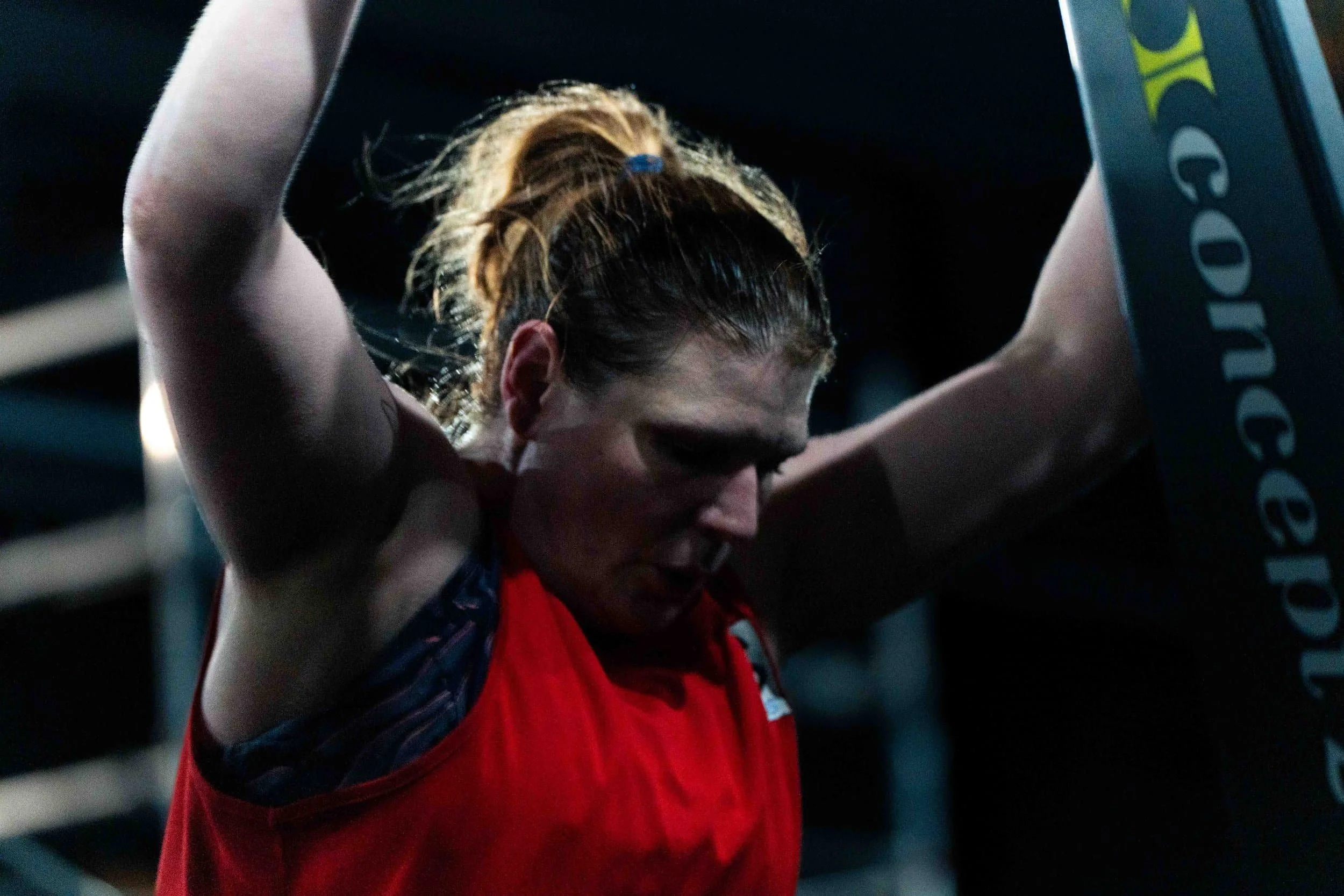 A woman in a red sports top is resting with her arms crossed above her head in a gym, appearing tired or focused.