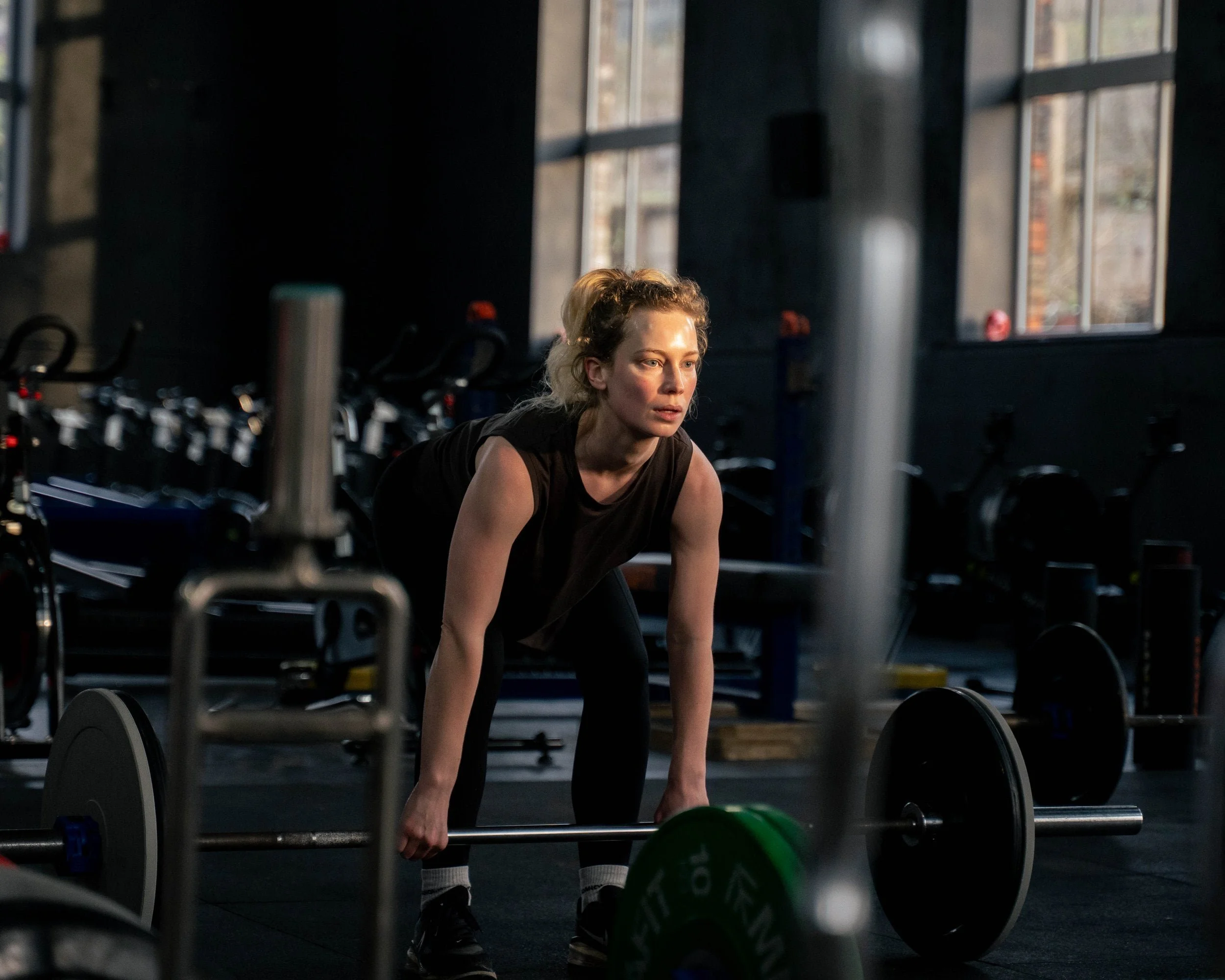 A woman in a black sleeveless top and black leggings is preparing to lift a barbell in a gym with black walls and large windows.