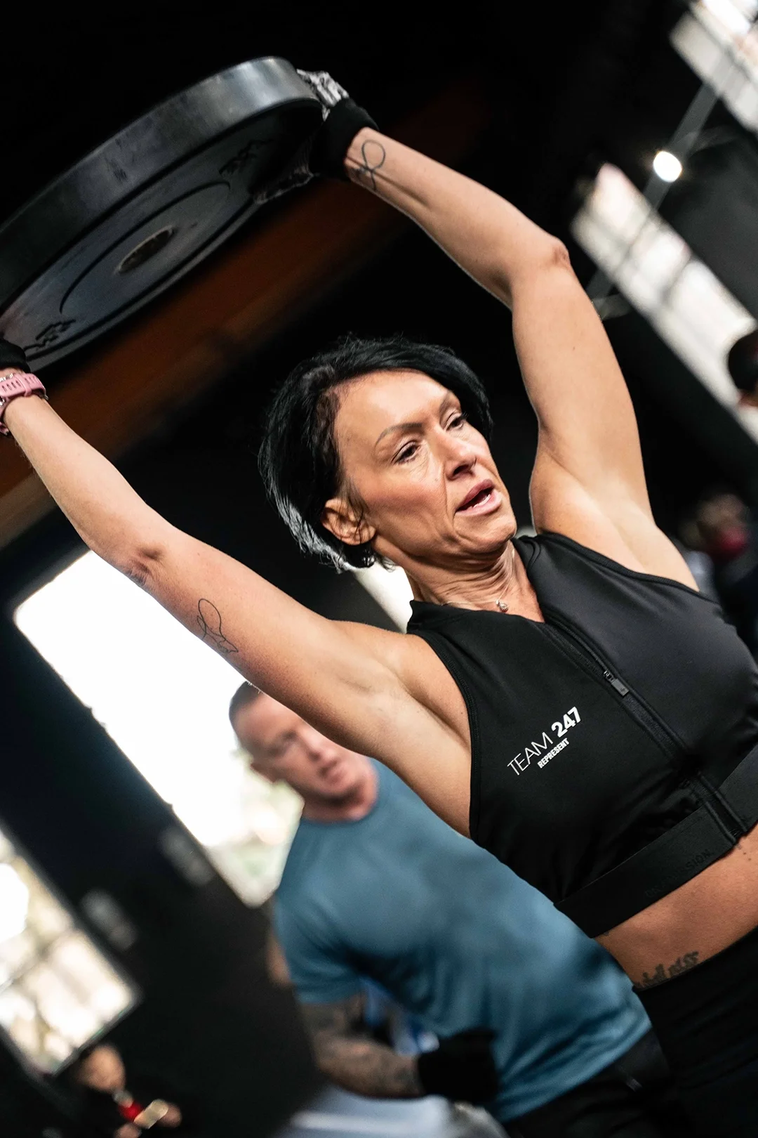 Woman in athletic wear lifts a weight plate overhead in a gym, with determination on her face. Other people are in the background.