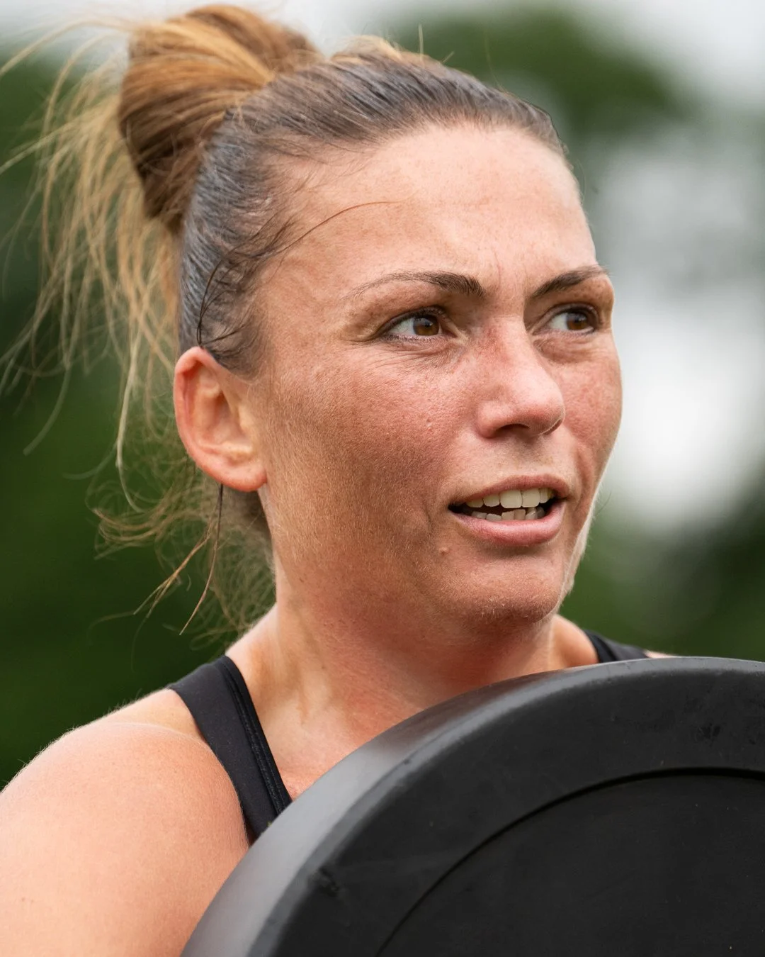 Woman with hair in a bun holds a weight plate outdoors, looking focused and slightly tired during the Deadly Dozen Dublin workout.