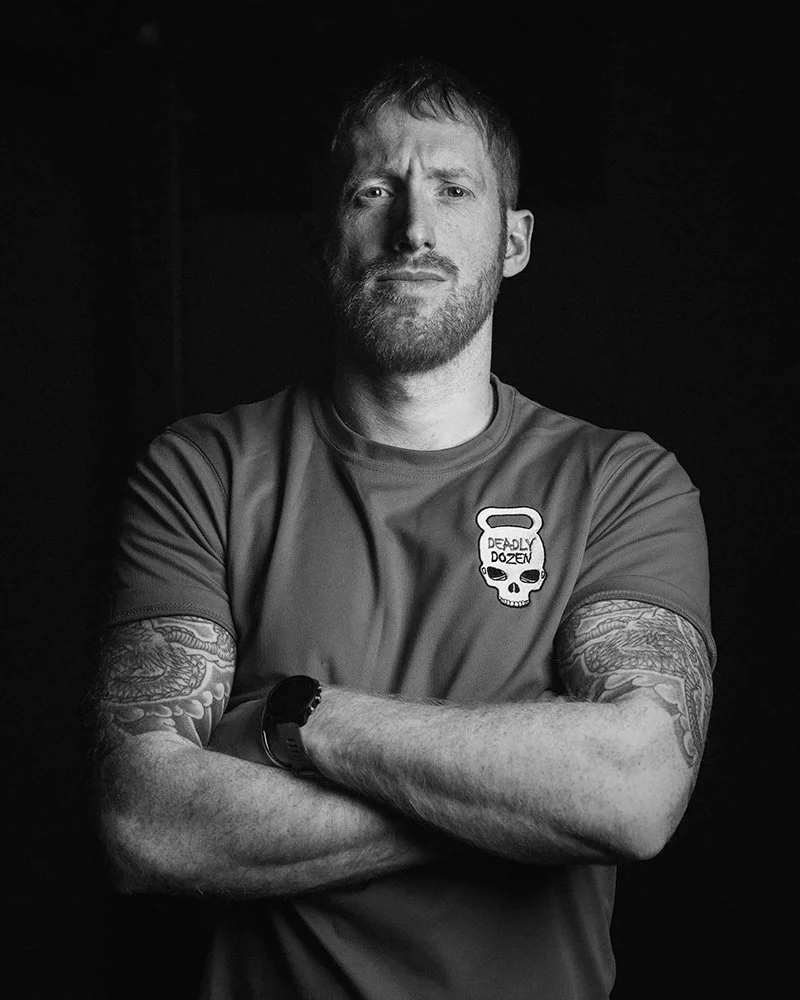 Jason Curtis, with tattoos and a serious look, stands arms crossed in a Deadly Dozen shirt against a dark background.