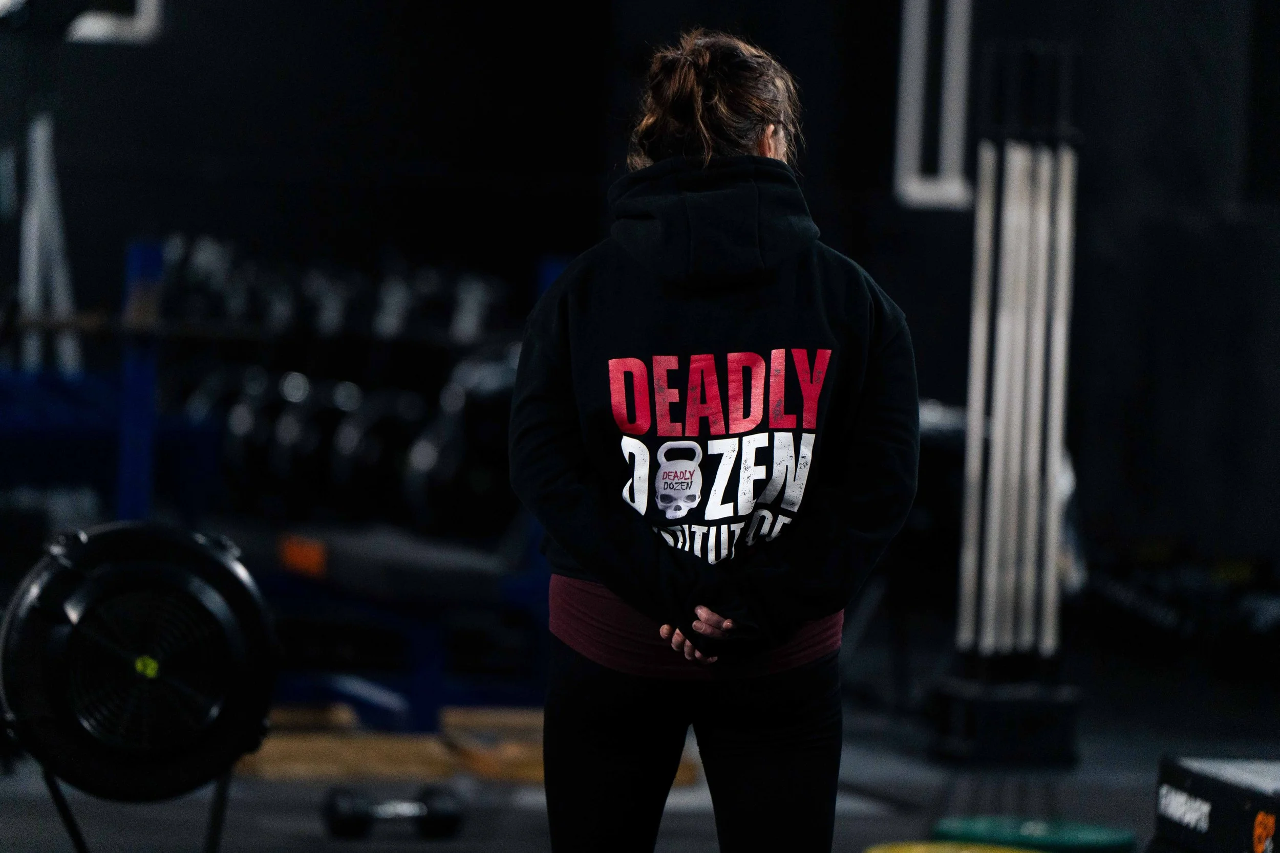 Person standing in a gym wearing a black hoodie with the words 'Deadly Dozen' and a skull graphic on the back, with gym equipment in the background.