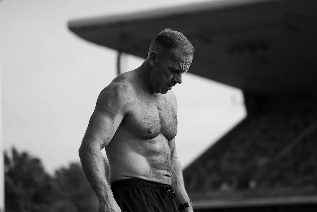 Black and white photo of a shirtless man with a muscular physique looking down, standing outdoors near what looks like a plane wing.