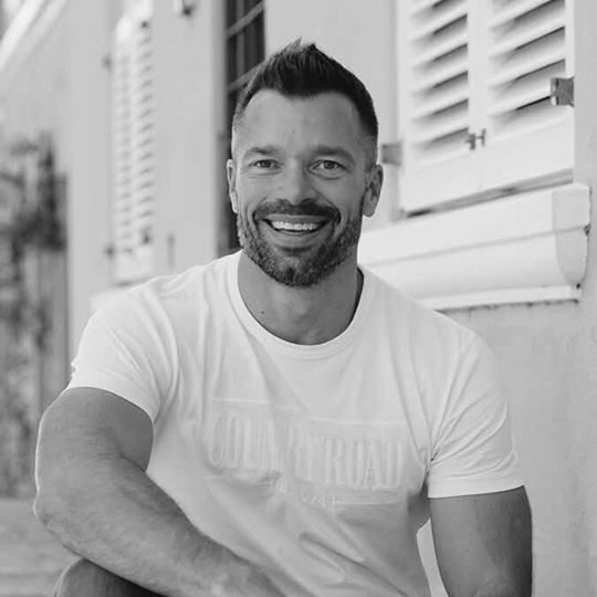 A smiling man with short dark hair and a beard, sitting outdoors near a building with shut window shutters, wearing a plain light-colored t-shirt.