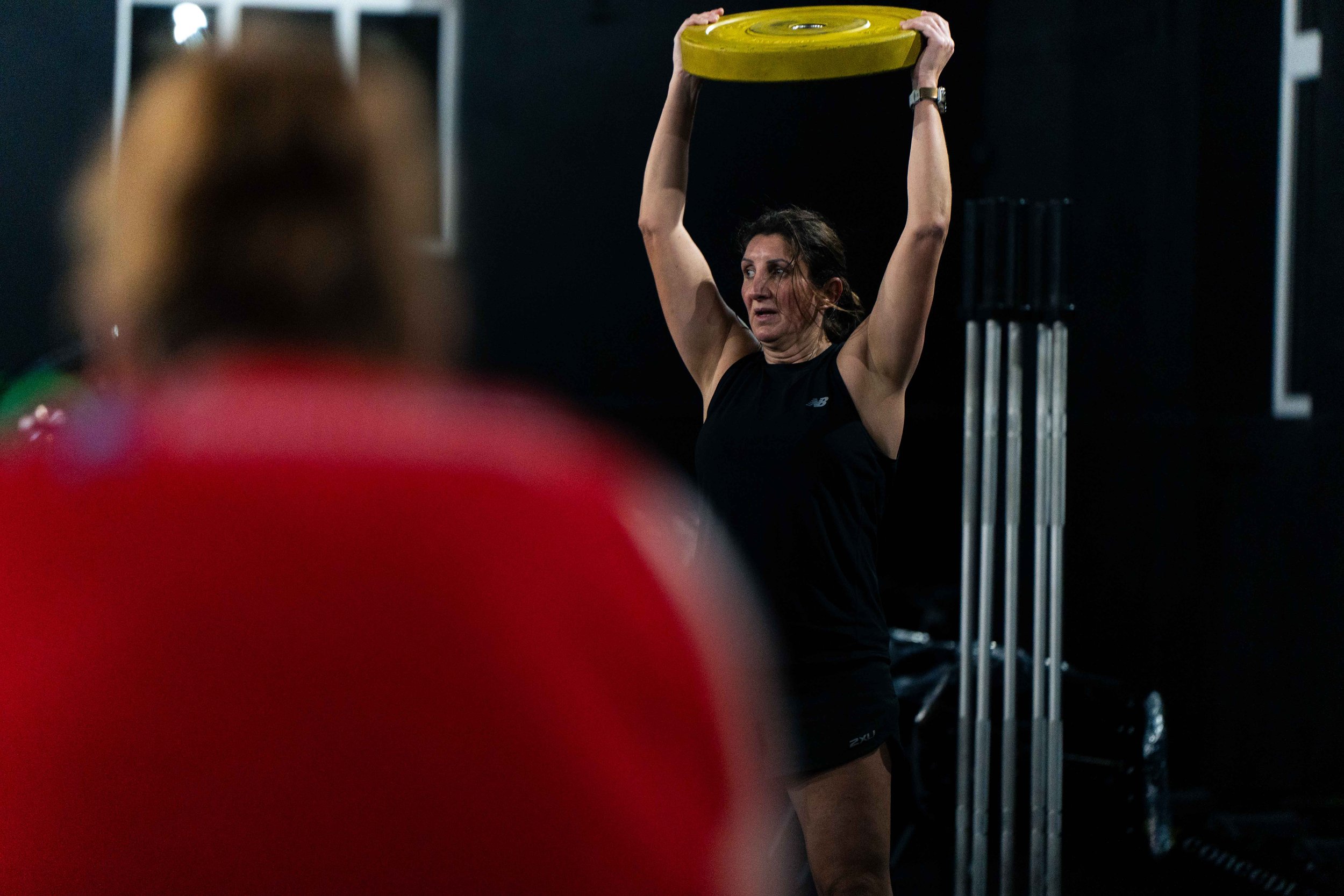 Woman lifting a yellow weight plate overhead in a gym, with a blurred figure in red in the foreground.