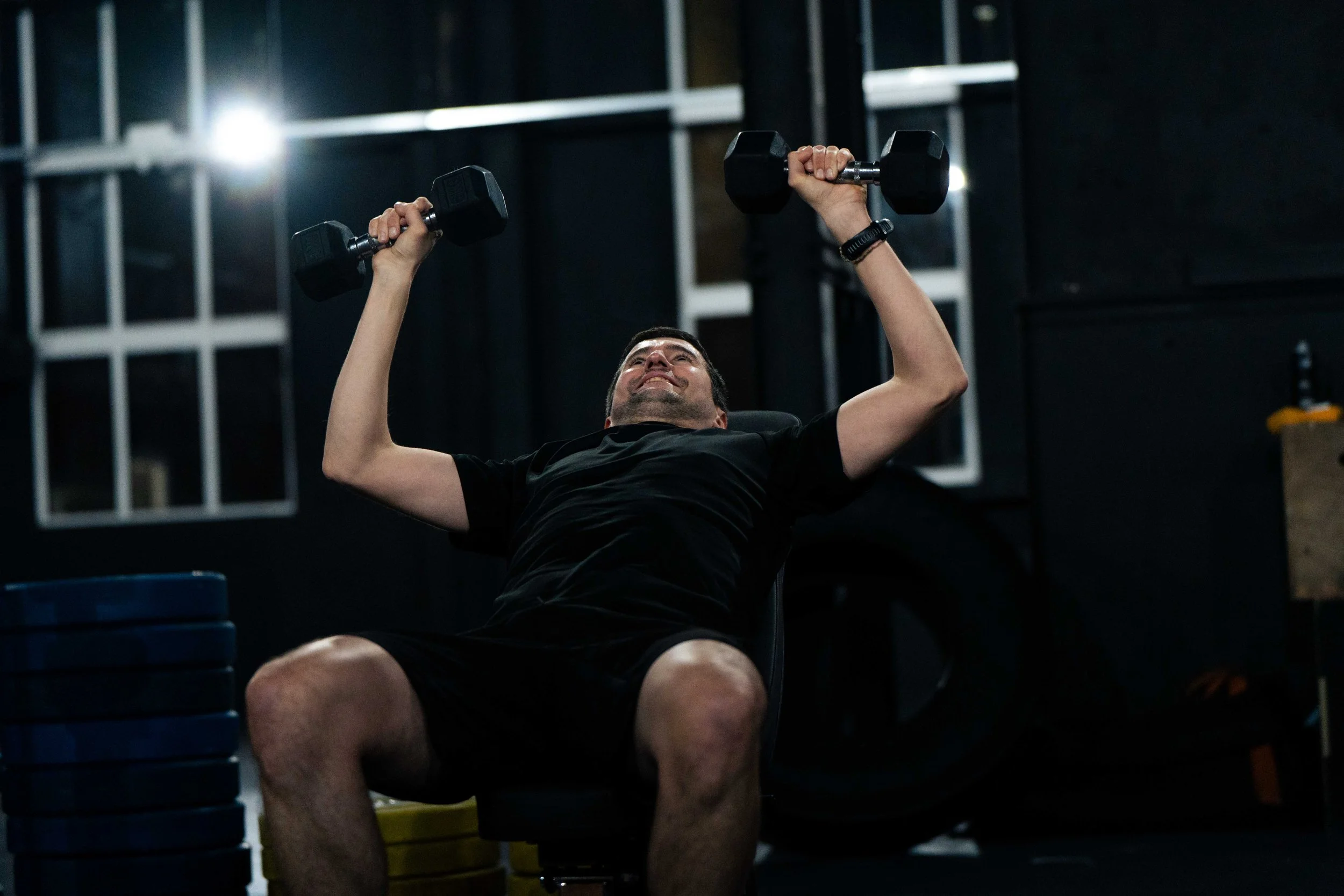 Man lifting dumbbells while seated in a gym, with black walls and equipment in the background.