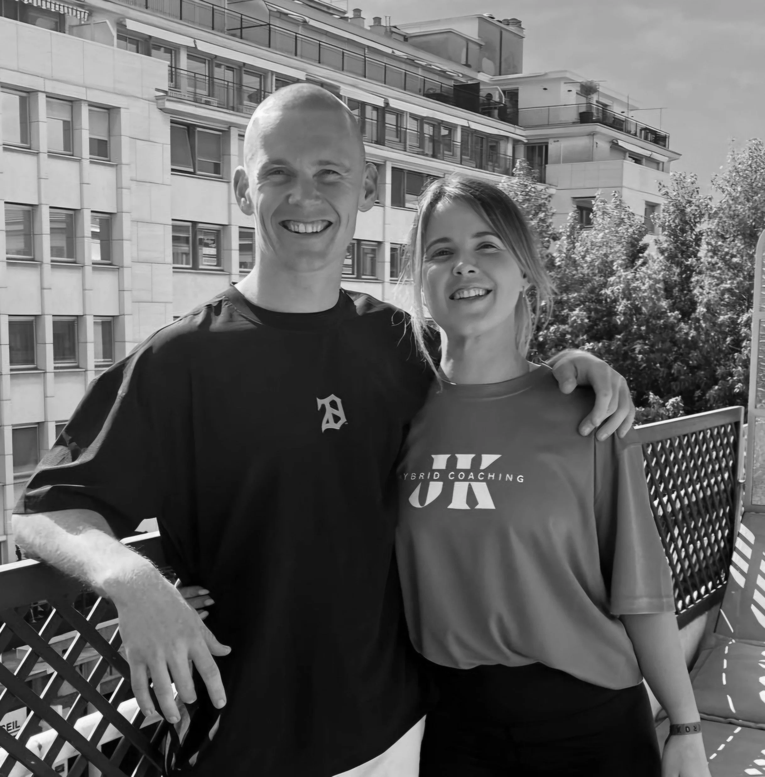 Two people smiling on a balcony, with urban buildings and trees in the background, in black and white.