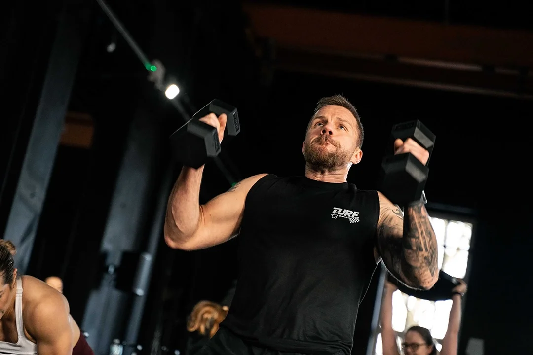 Man wearing a black tank top lifts dumbbells in a gym, focused and exerting effort during his workout.