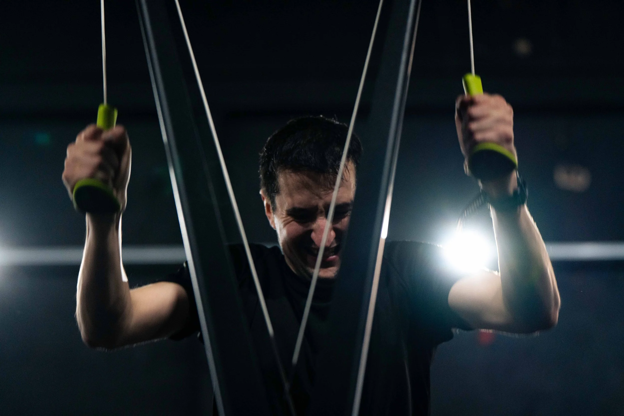 A man with short dark hair is lifting a weighted fitness machine in a gym, with visible strain and effort on his face. Bright light is shining behind him, creating a backlit effect.