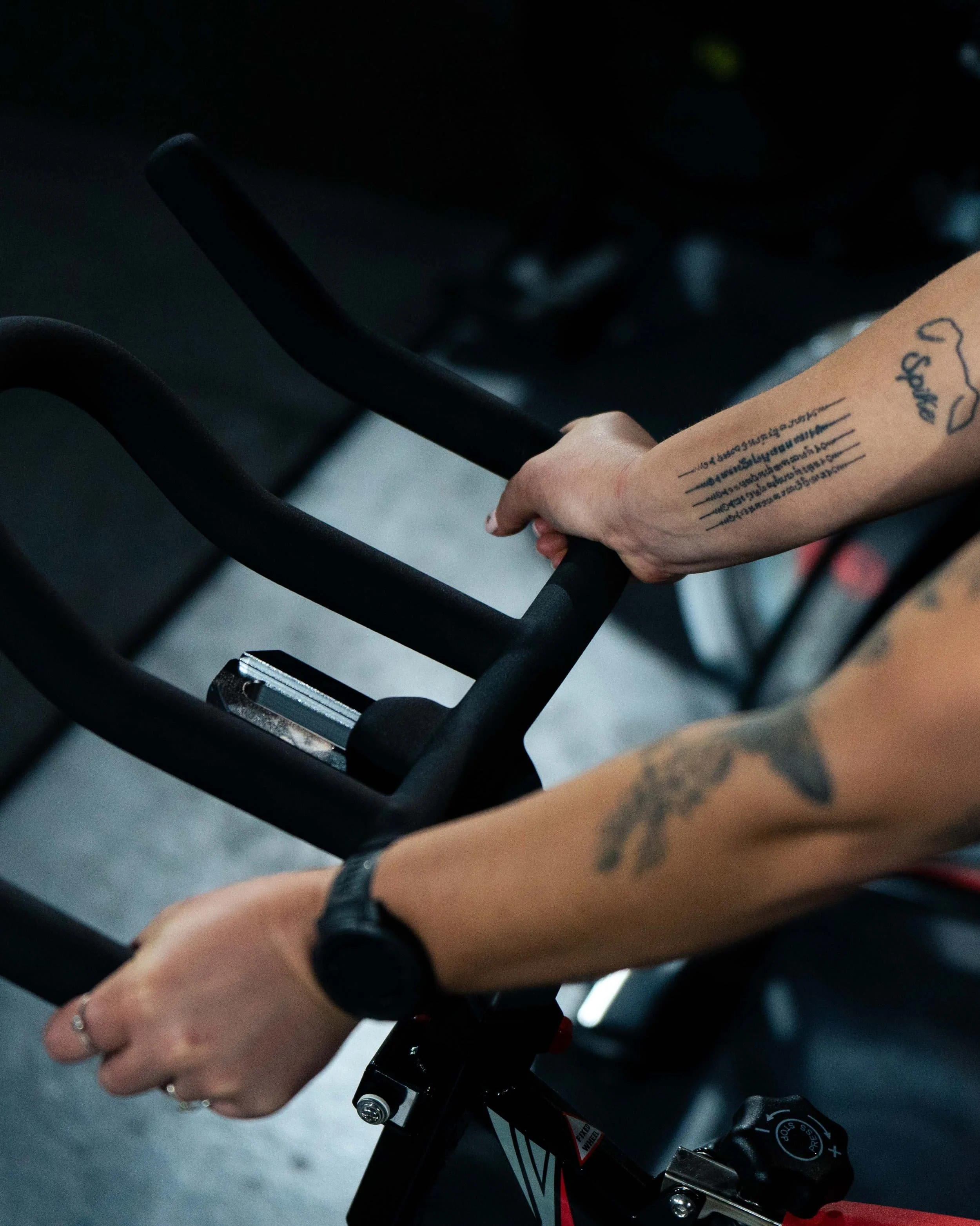 Close-up of a person’s hands holding the handlebars of a stationary exercise bike in a gym, with visible tattoos and a black smartwatch.