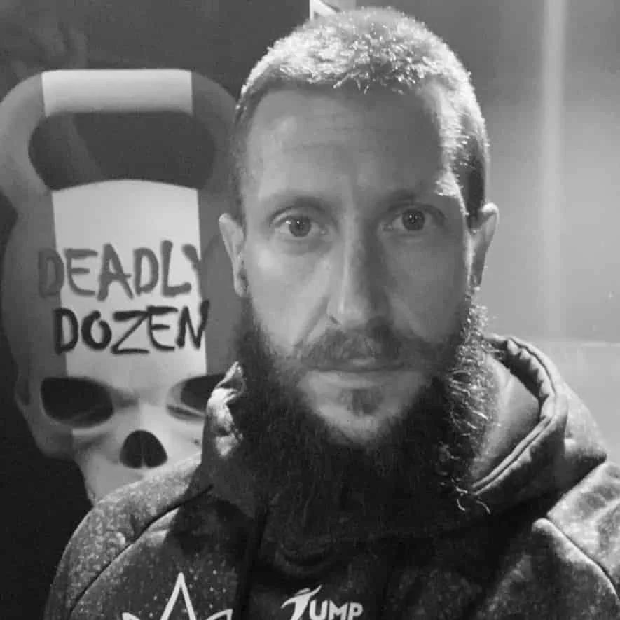 A man with a beard and short hair taking a selfie in front of a poster for the video game Deadly Dozen, which features a skull and the game's title. The photo is in black and white.