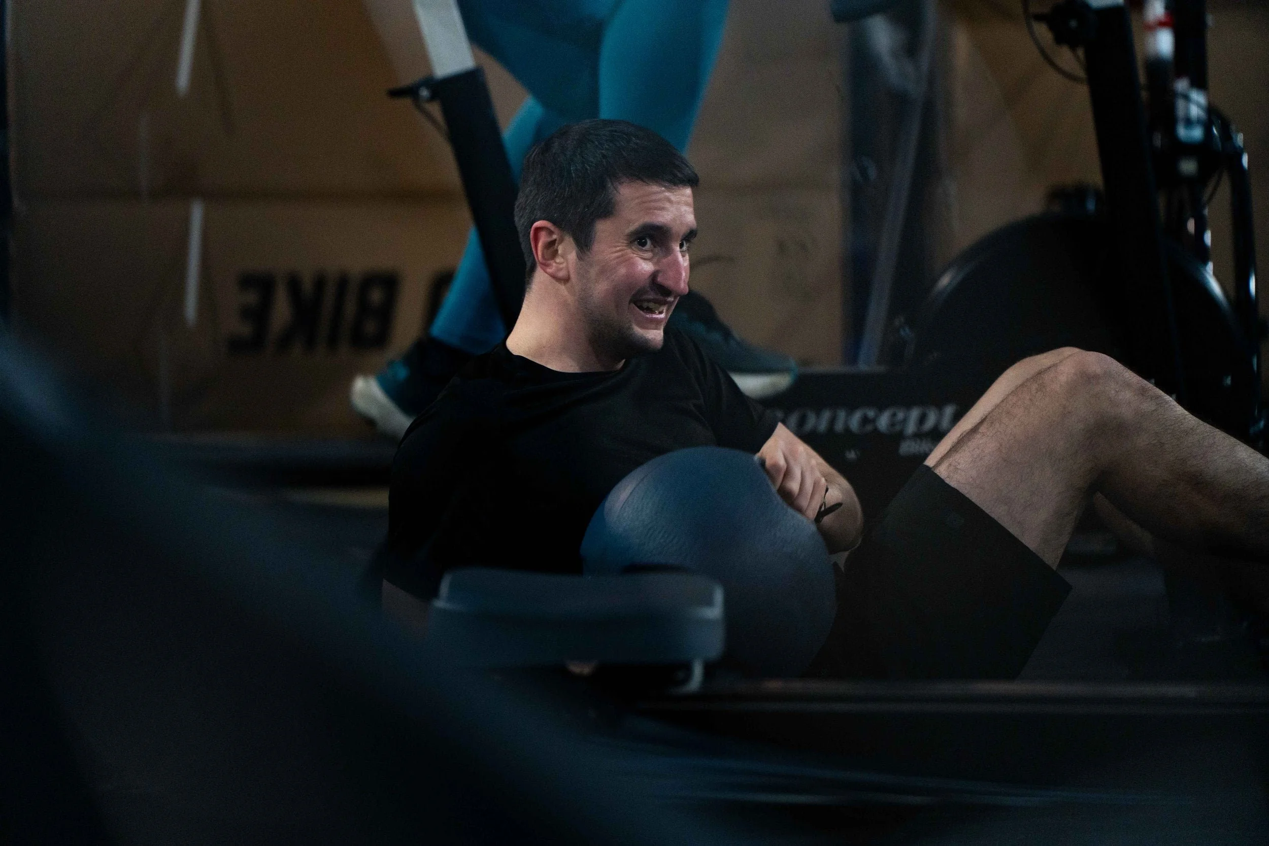A man exercising on the floor of a gym, holding a medicine ball and smiling